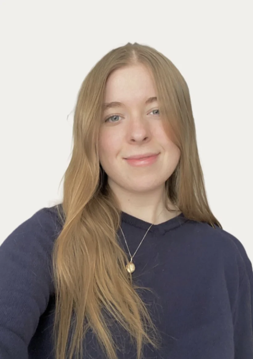 A young woman with long, wavy red hair and blue eyes, smiling slightly, wearing a navy blue sweater and a delicate necklace with a pendant, standing against a plain white background.
