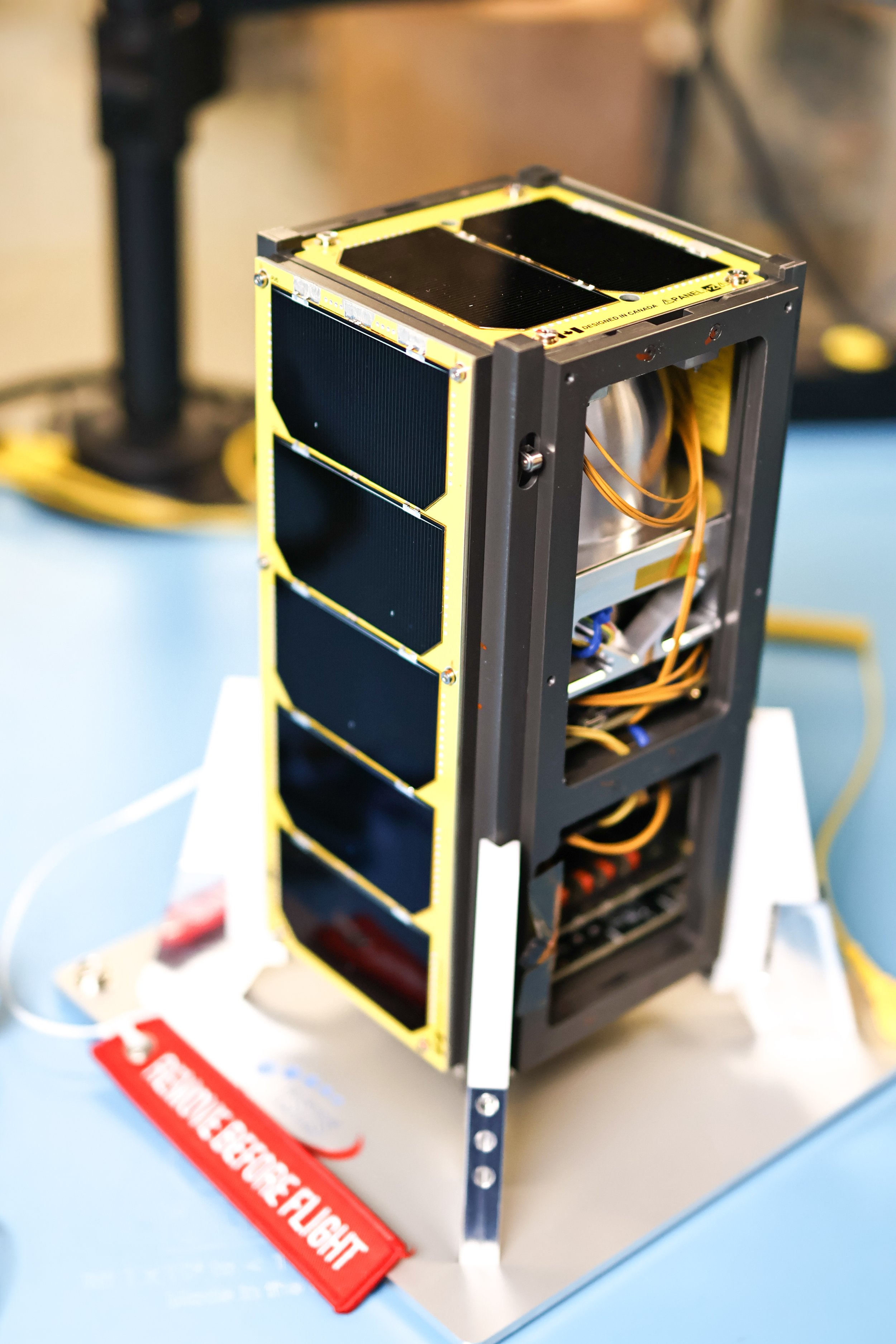Small satellite with black solar panels mounted on a yellow frame, placed on a white surface with a red fire extinguisher tag nearby.