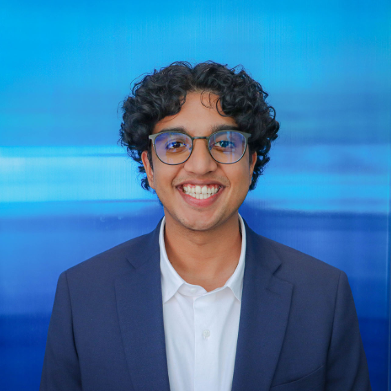 A young man with curly black hair, glasses, and a wide smile, wearing a navy blazer and white shirt, standing in front of a blue gradient background.