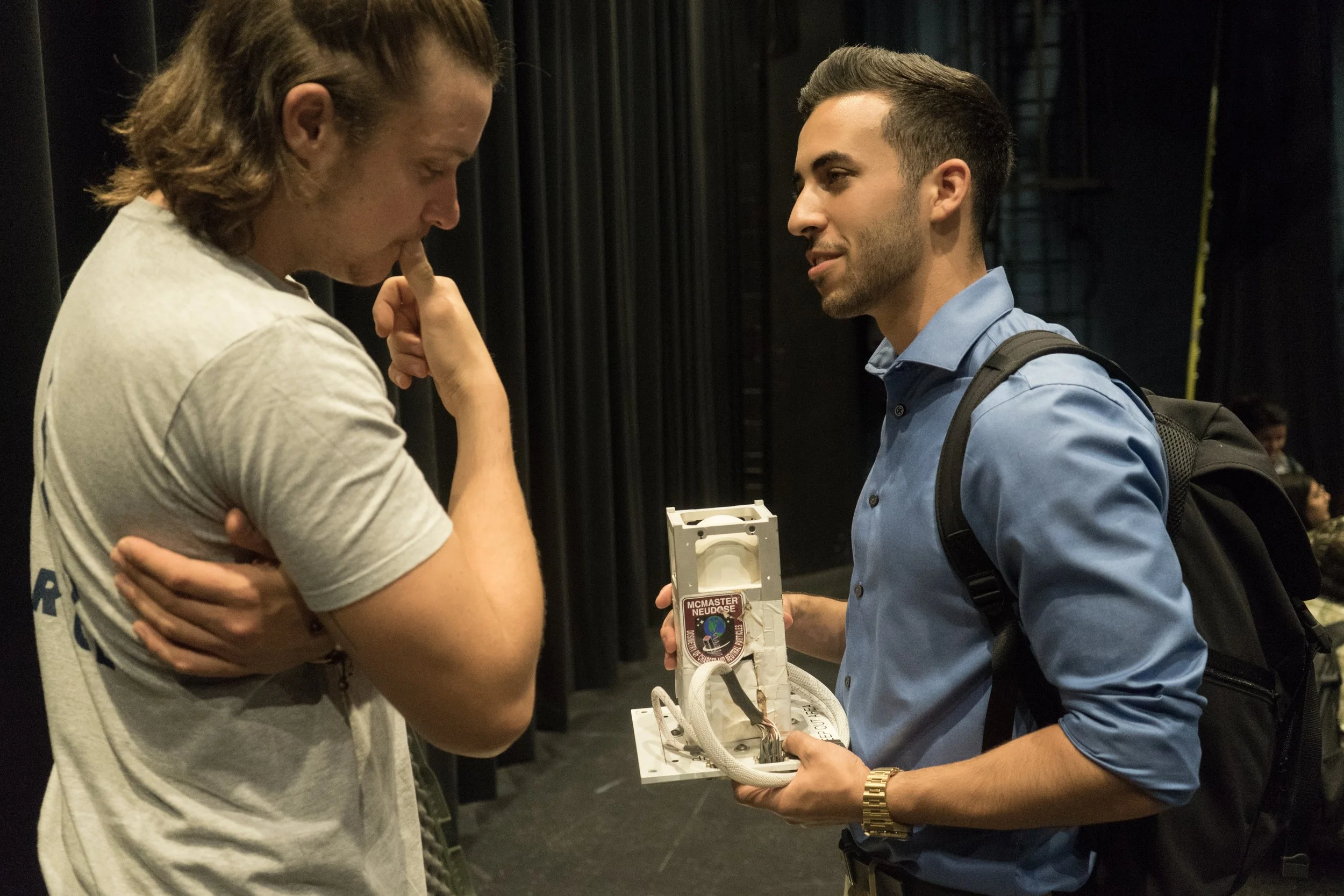 Two young men engaged in a conversation backstage, one holding a device with wires and a sticker, wearing a backpack, and the other with a thoughtful expression, wearing a casual t-shirt.