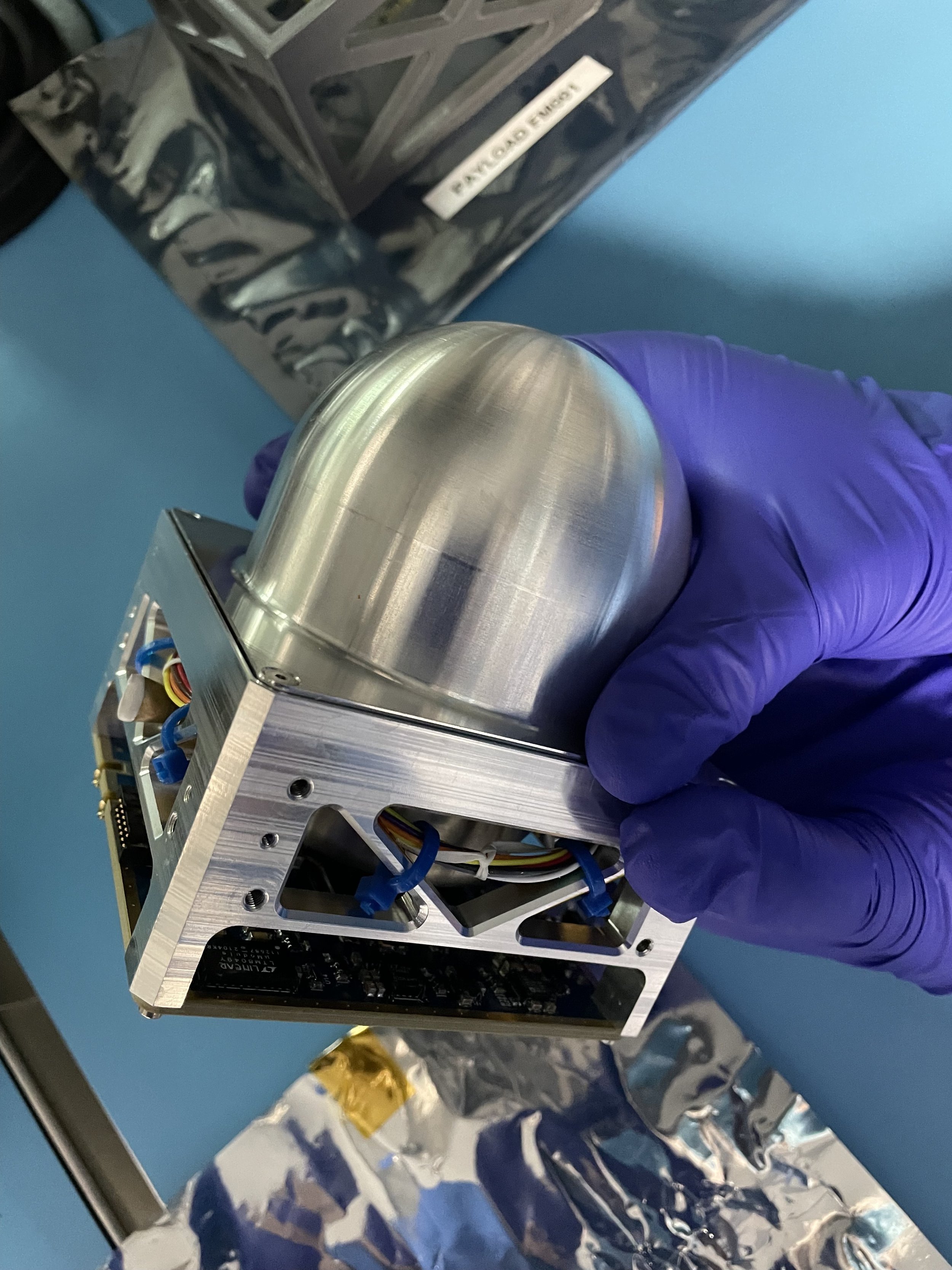 Person wearing purple gloves holding a metallic, dome-shaped object integrated with a circuit board assembly, on a blue surface with electronic components and packaging materials nearby.