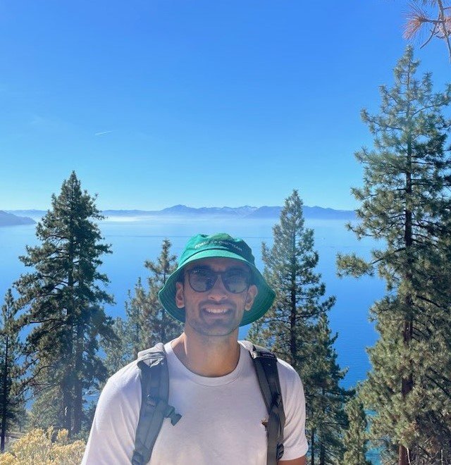 Smiling man with sunglasses and a green hat, standing outdoors with pine trees and a lake in the background under a clear blue sky.
