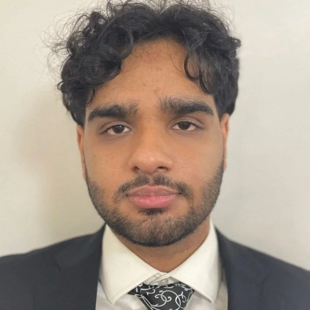 A young man with dark, curly hair, a beard, and a mustache wearing a black suit, white shirt, and a patterned tie, posing against a plain light-colored background.