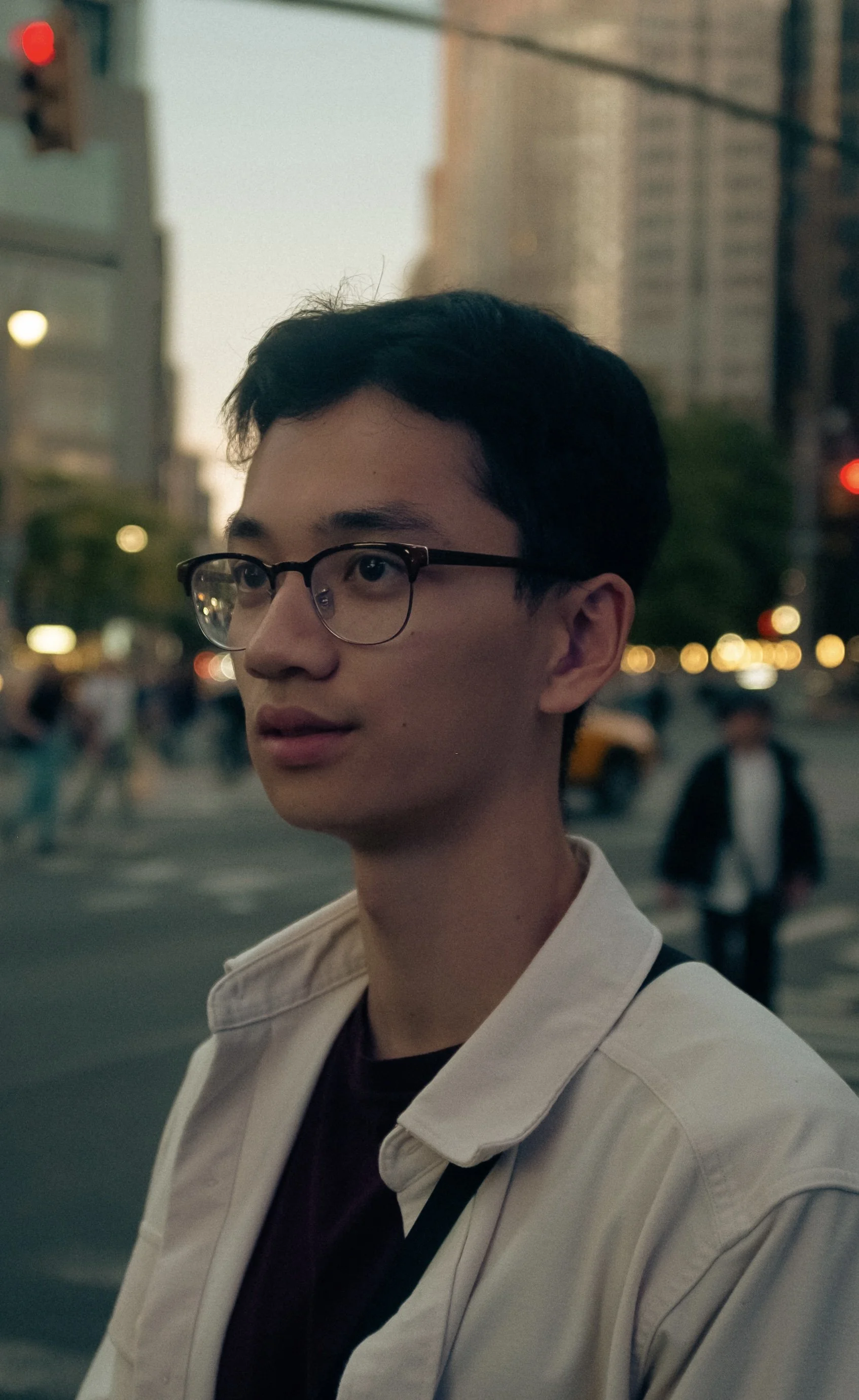 A young man with glasses wearing a white jacket standing on a city street during dusk, with blurred city lights and buildings in the background.