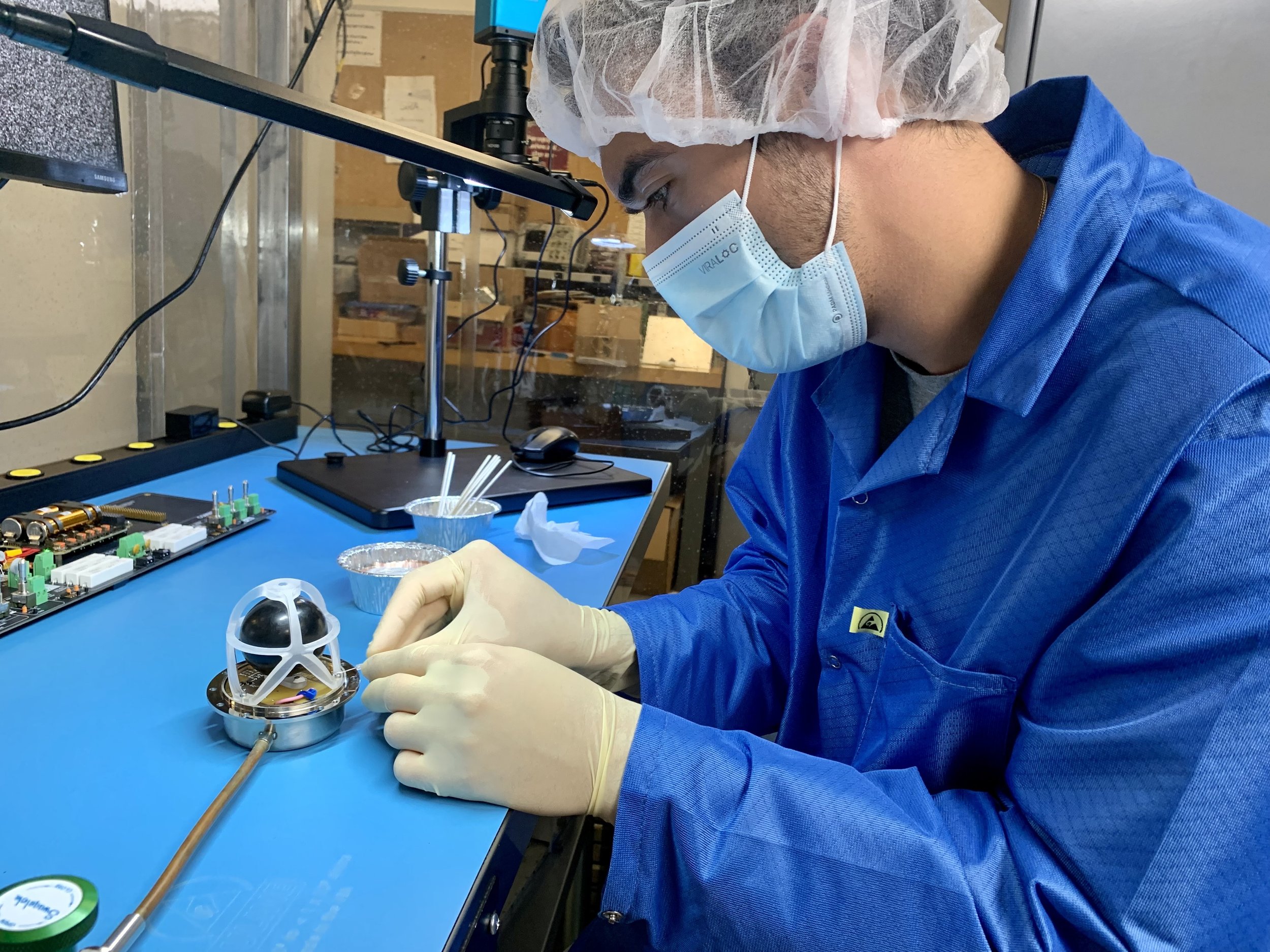 A scientist wearing a blue protective coat, a hair cap, a face mask, and gloves is working on a small robot in a laboratory. The lab has electronic components, a microscope, and tools on the workbench.