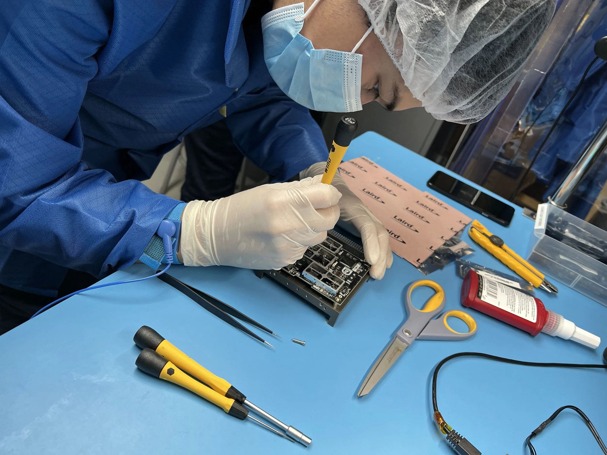 A technician working on an electronic circuit board at a workbench with various tools including screwdrivers, scissors, and a glue tube, wearing gloves, a face mask, and a hair cover.