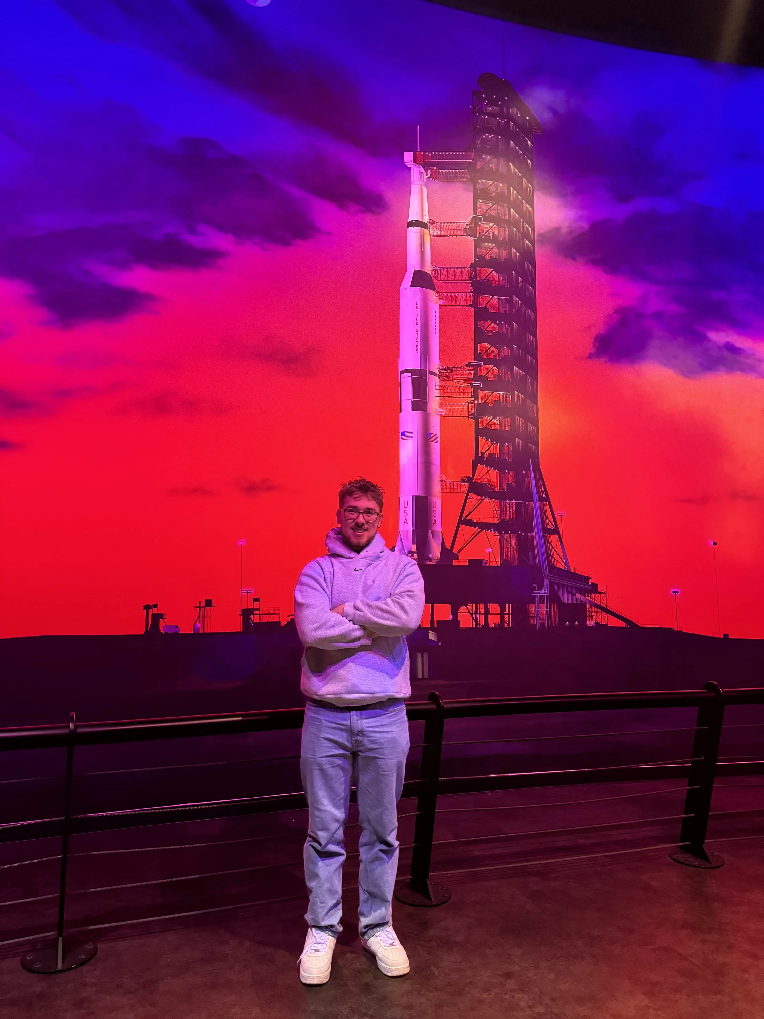 A person smiling and standing in front of a large digital display of a rocket on a launch pad against a colorful sky background at an exhibit or museum.