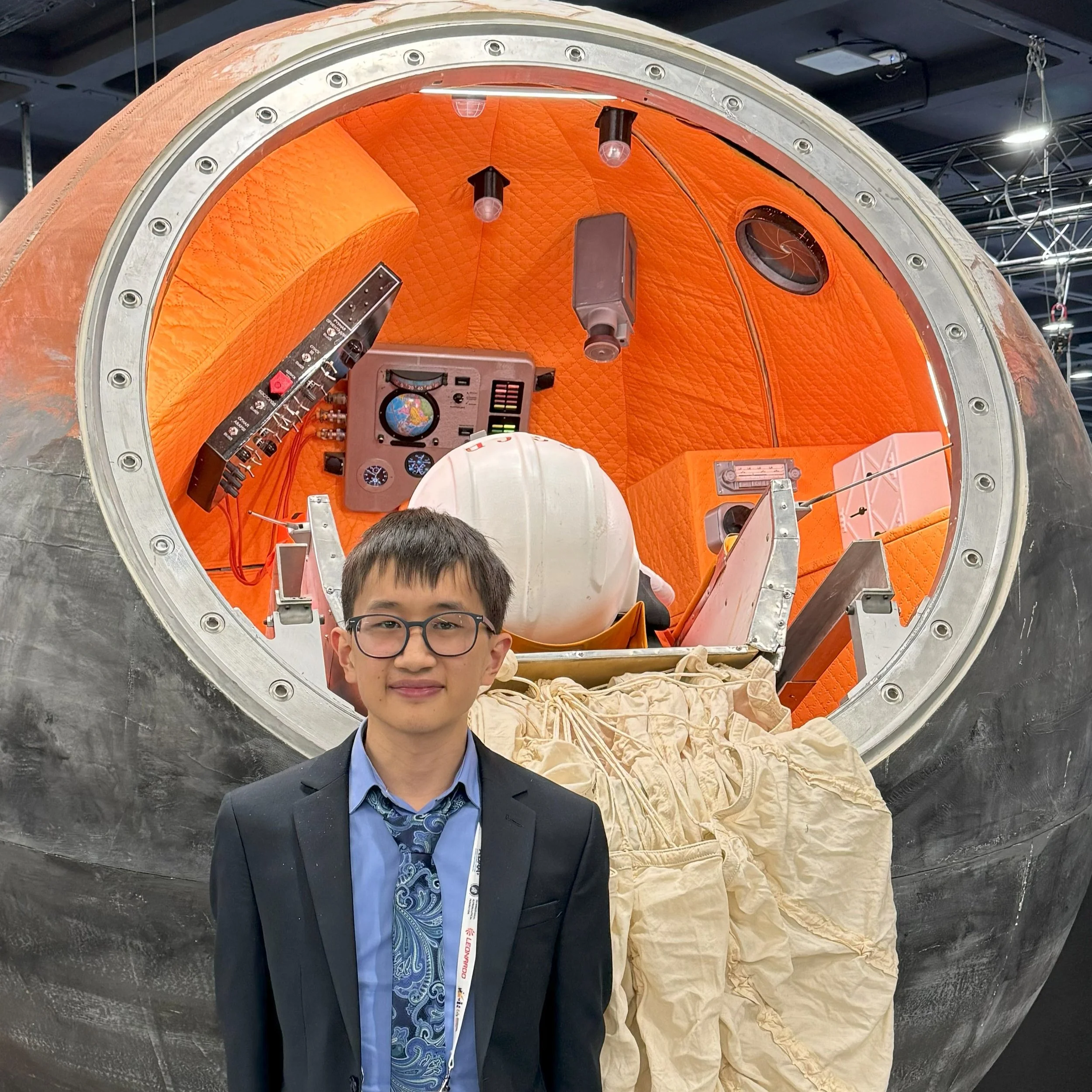 A young boy in glasses and a suit stands in front of a space capsule mock-up with an orange interior, containing various control panels, instruments, and a space suit.