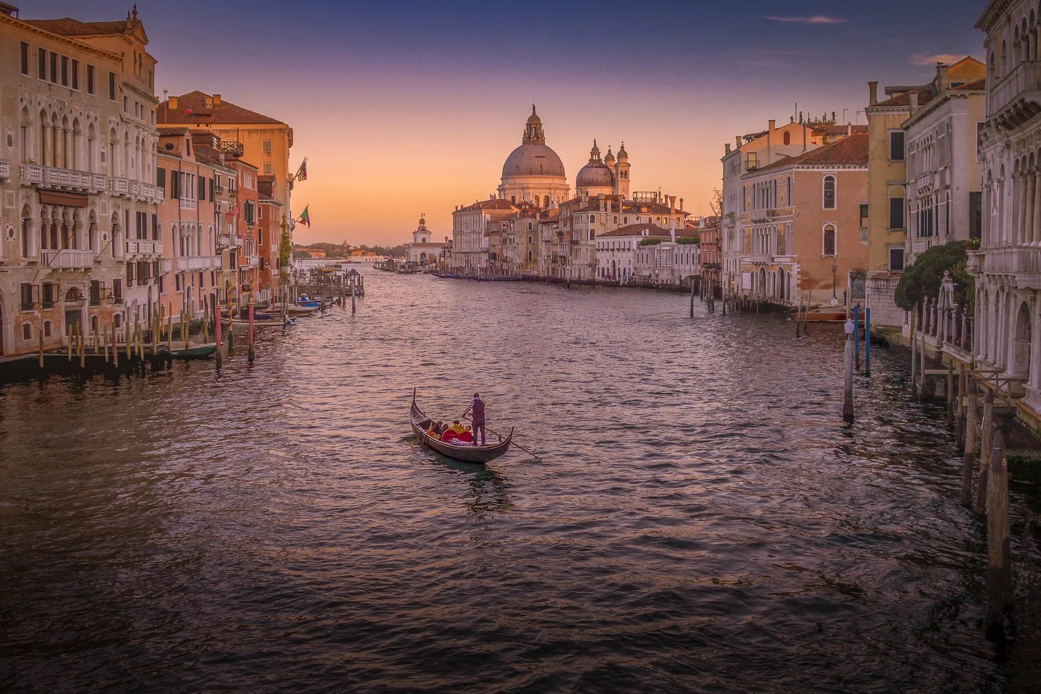 View of a canal in Venice, Italy, during sunset with gondola in the foreground and historic buildings along the sides of the canal, culminating with the domes of a church in the background.