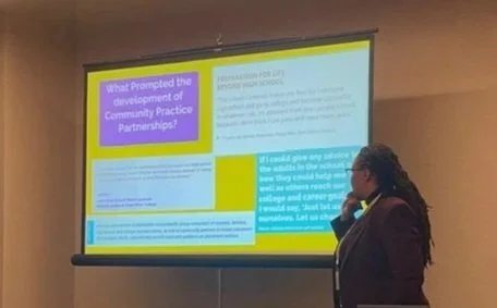 A woman standing in front of a presentation screen displaying slides on a wall. The slides are titled 'What Prompted the development of Community Practice Partnerships?' and contain text and colorful sections in purple, yellow, and blue.