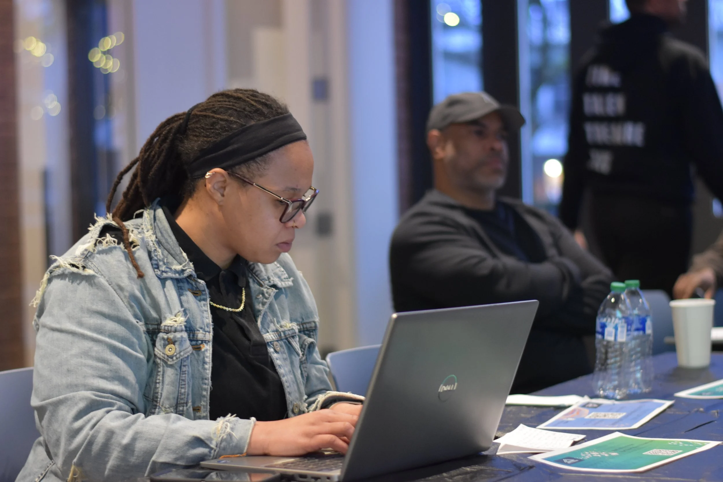A woman with glasses and dreadlocks working on a laptop at a table, with a man sitting behind her at the same table. The scene appears to be indoors, with several papers, water bottles, and a paper cup on the table.