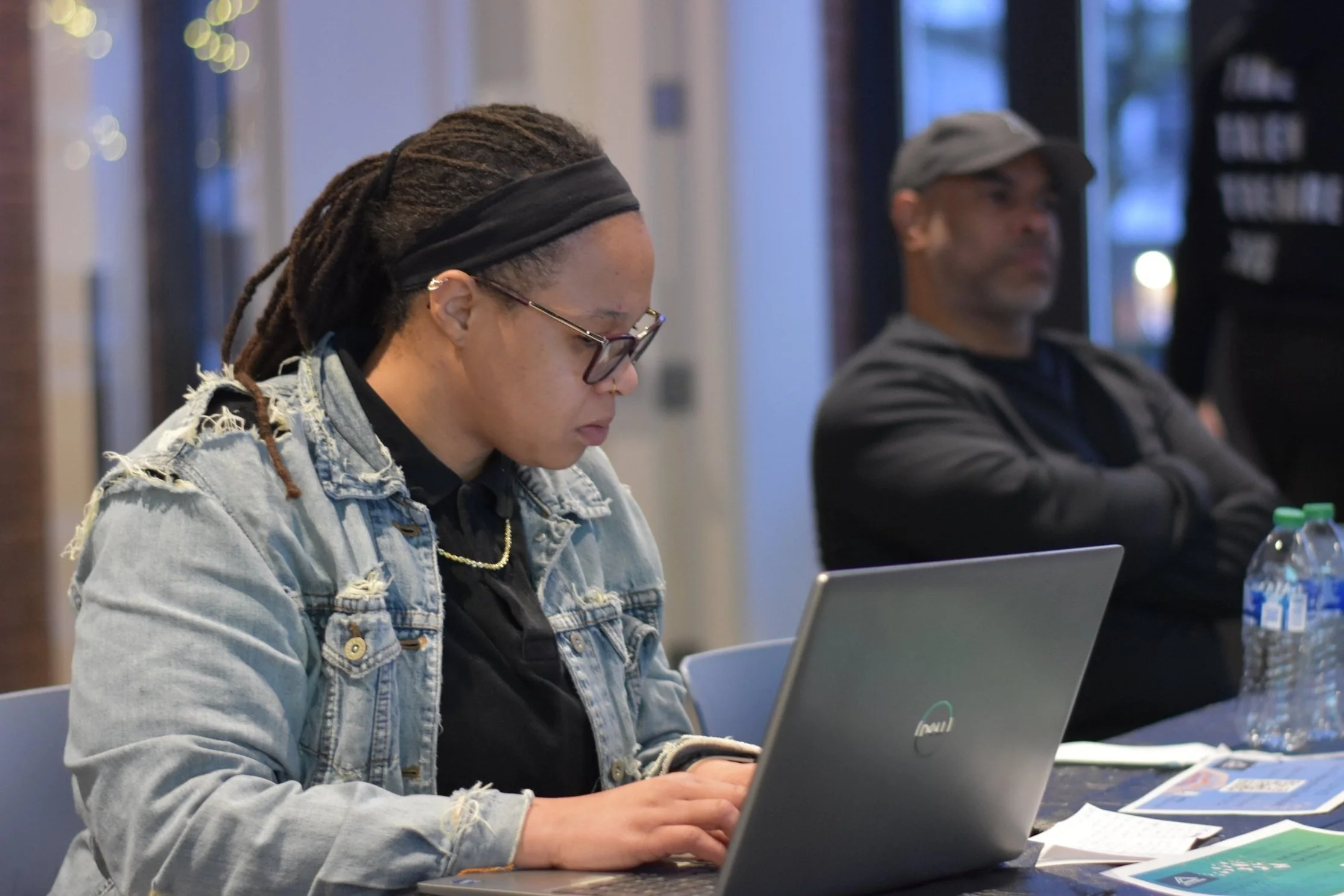 A woman with glasses typing on a laptop at a table, with a man sitting with arms crossed in the background, inside a room with windows.