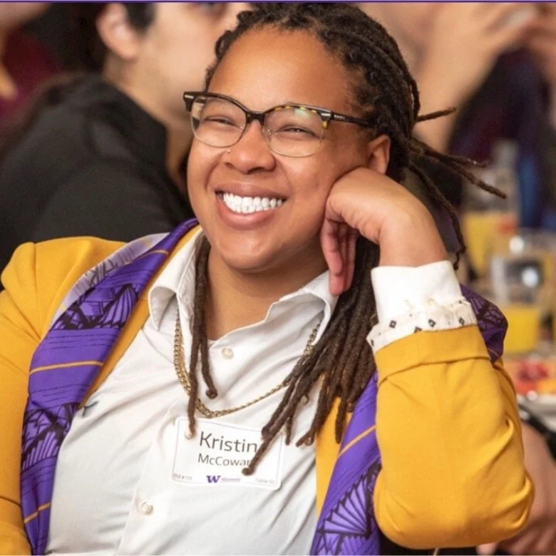 A woman with glasses and dreadlocks smiling and resting her head on her hand, wearing a white shirt and yellow blazer.
