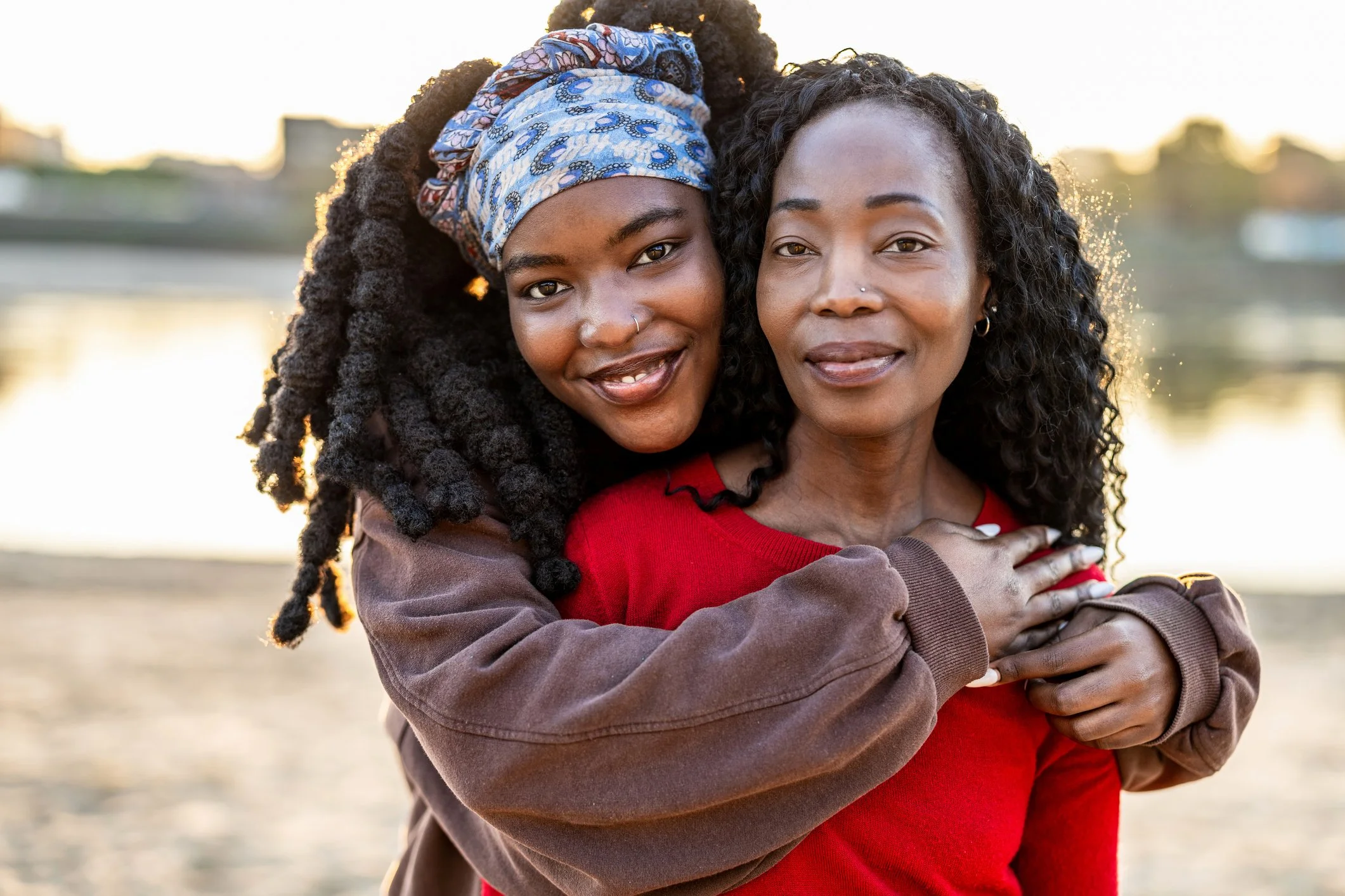 Two women hugging and smiling by the water at sunset, one in a patterned headwrap.