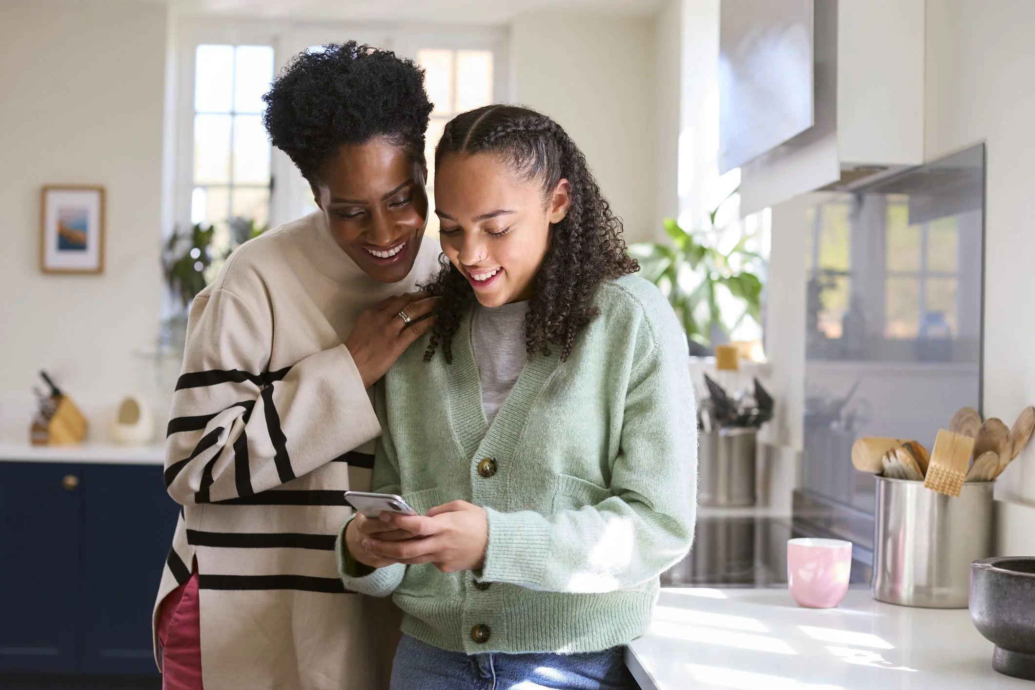 A mother and her teenage daughter smiling together over a phone in a kitchen.