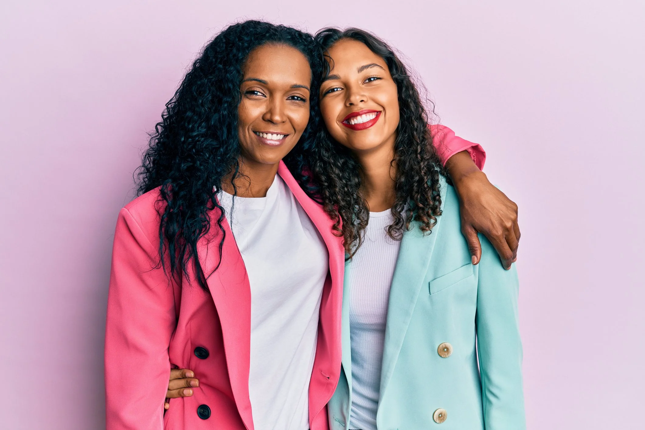 Two women in pink and teal blazers smiling with their arms around each other.