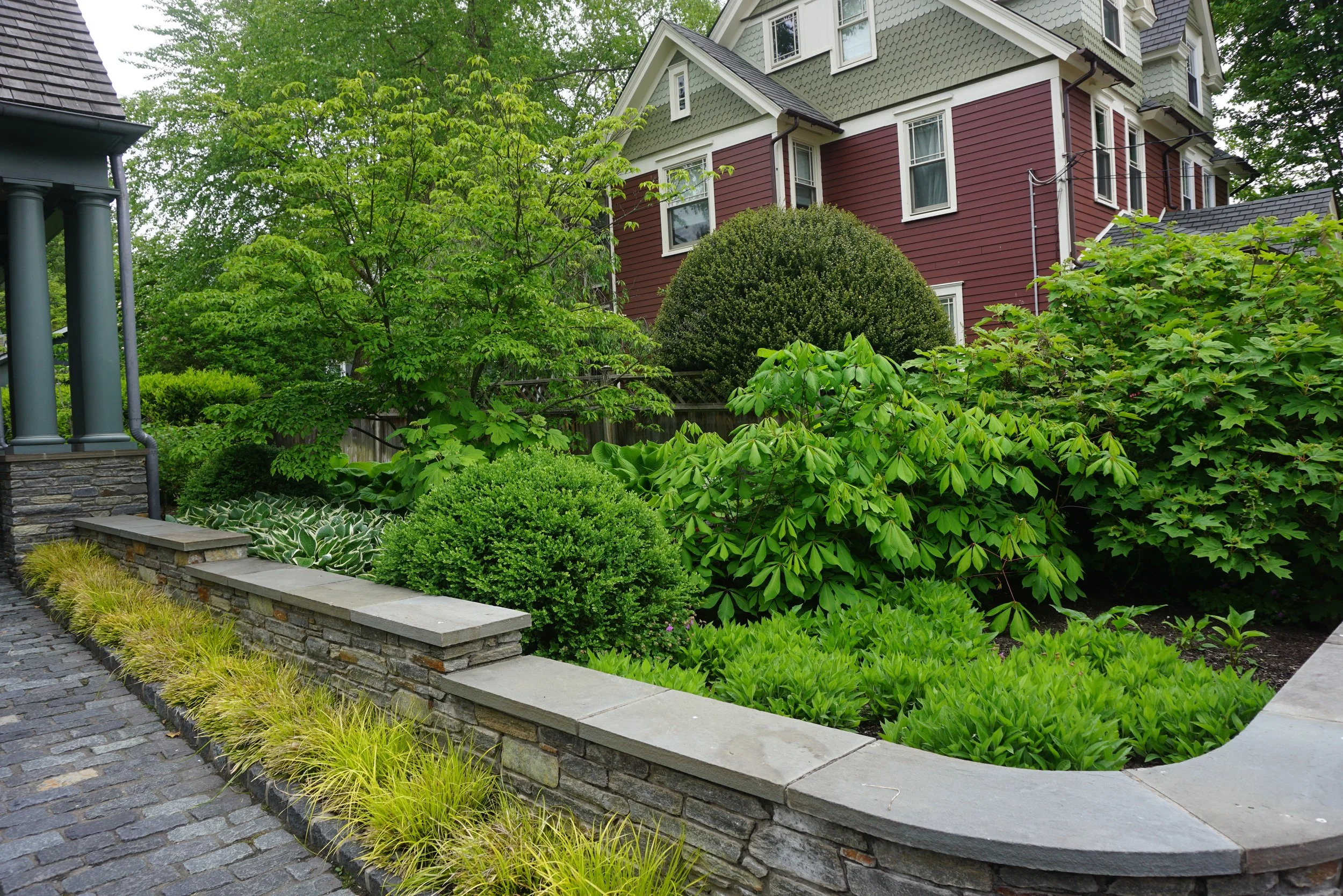 A garden with lush green bushes and trees next to a stone patio in front of a red house with white trim.