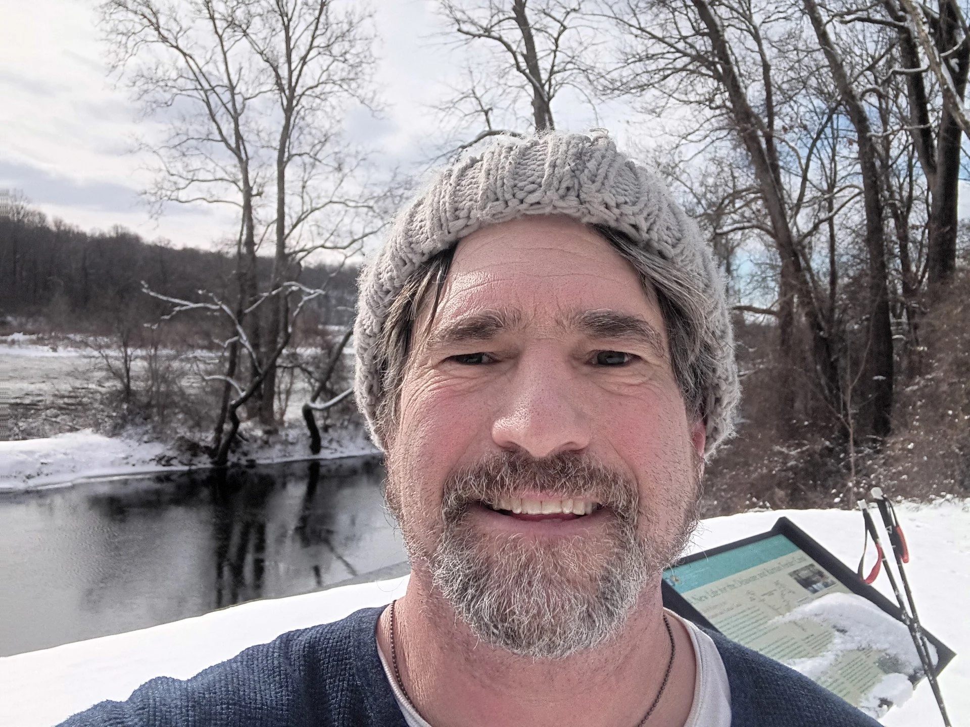 A man is smiling outdoors in winter, wearing a knit hat, with a snowy landscape and a river behind him.