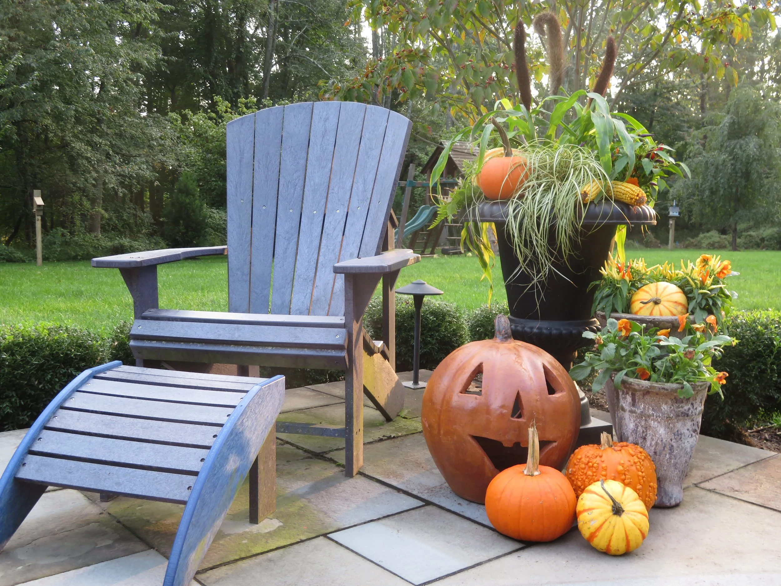 Outdoor patio scene with a blue wooden chair and footrest, pumpkins, and large potted plants with fall decorations in a backyard garden.