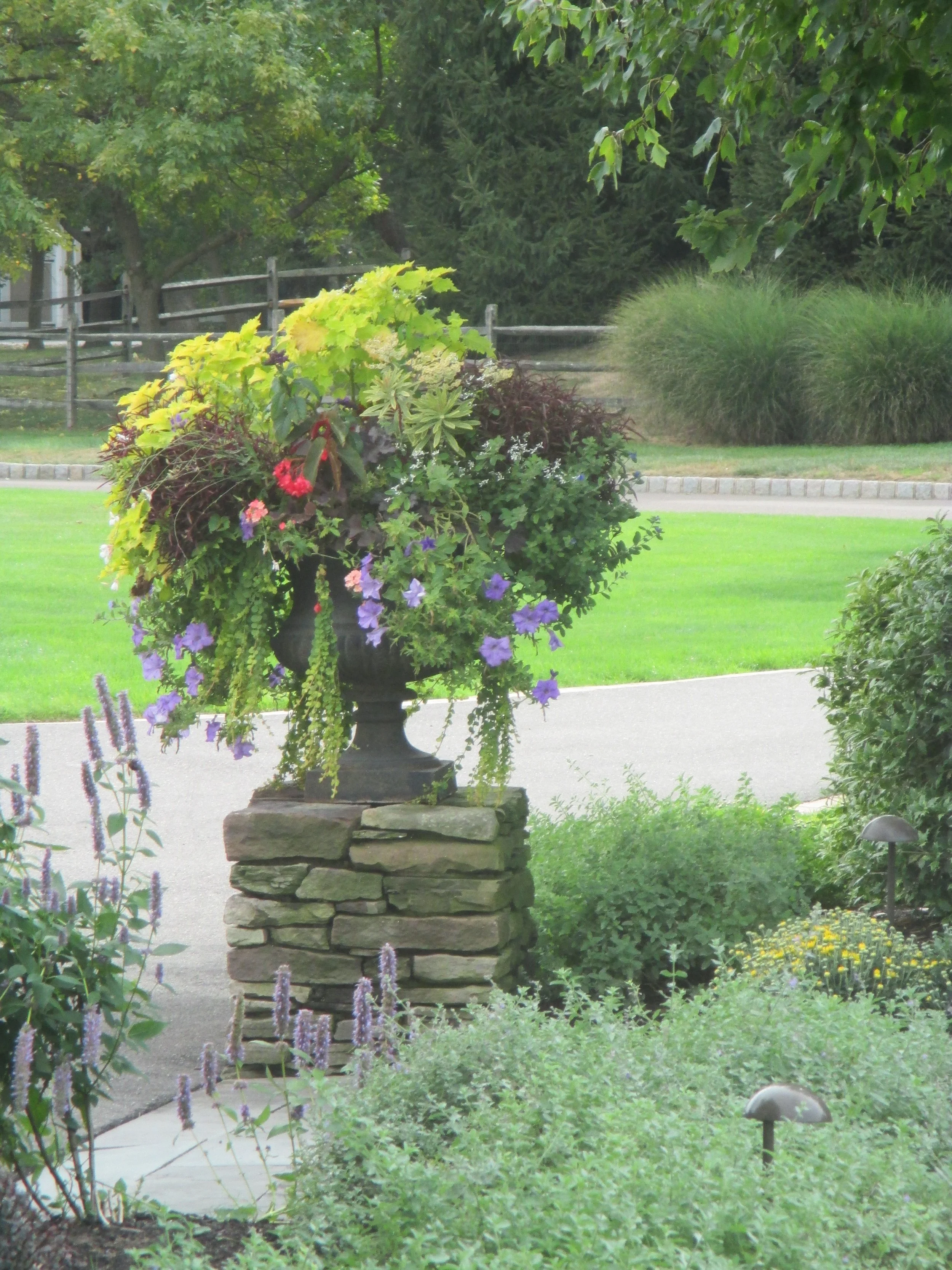A decorative stone pillar with a large urn filled with colorful flowers and greenery, situated in a landscaped garden with a lawn and trees in the background.