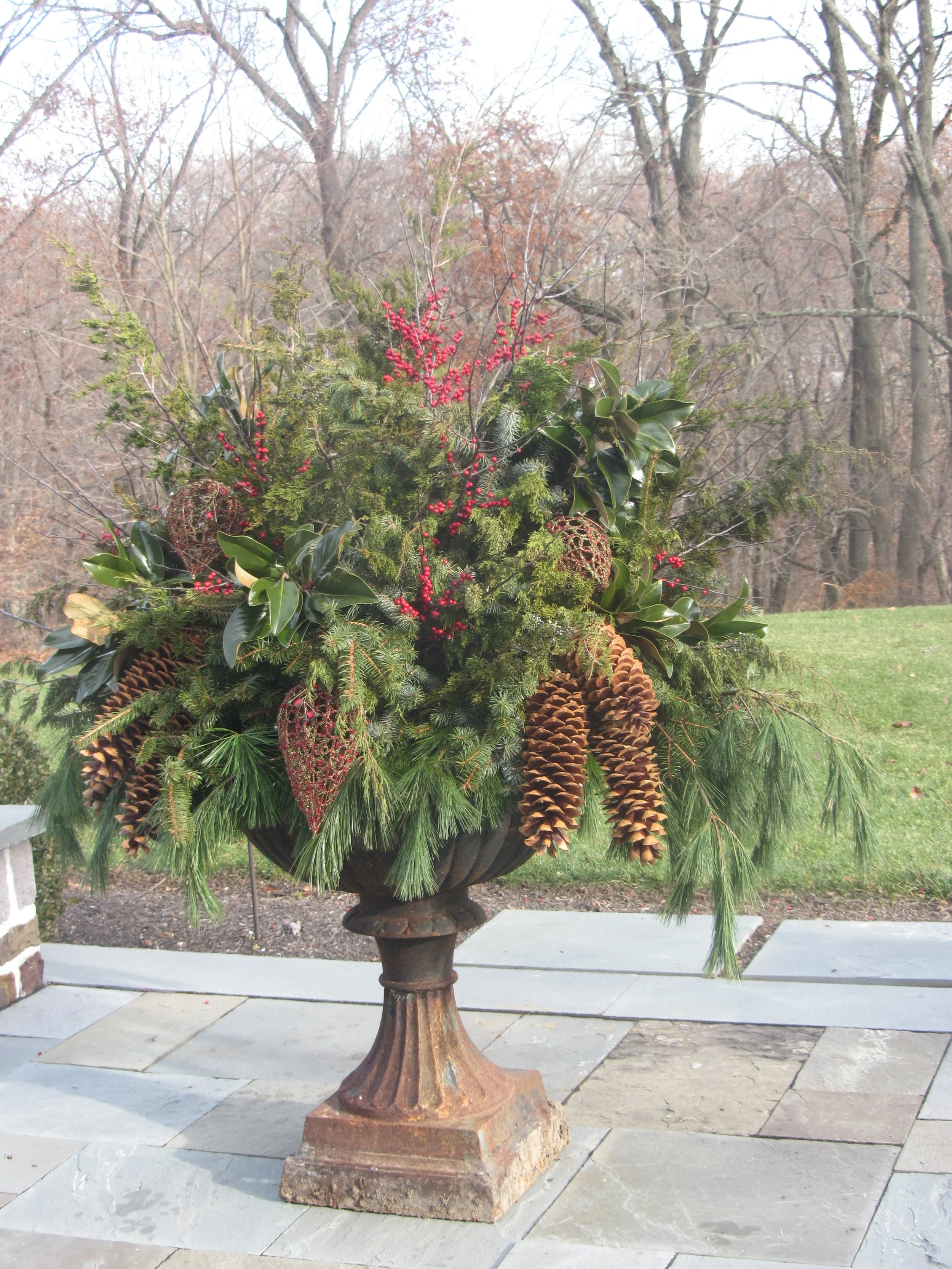 A large, decorative outdoor floral arrangement in an urn-shaped planter on a stone patio, with greenery, pinecones, red berries, and various plants amidst trees with bare branches in the background.