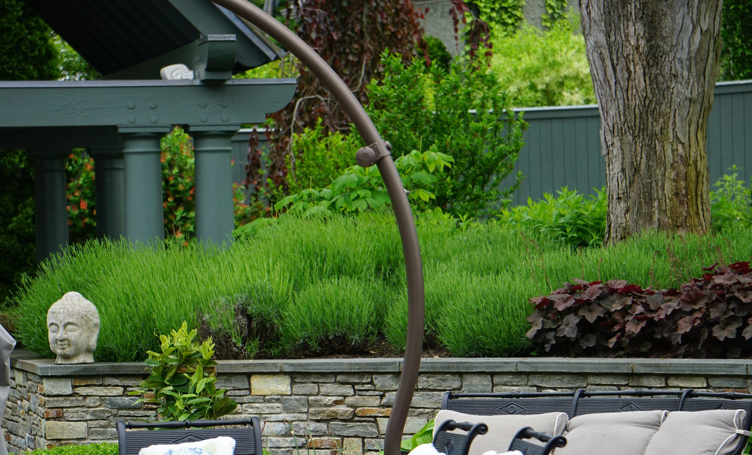 Backyard garden with a stone retaining wall, lush green plants, a decorative stone Buddha head, a small shrub, a large tree, outdoor furniture, and a garden heater or stand.