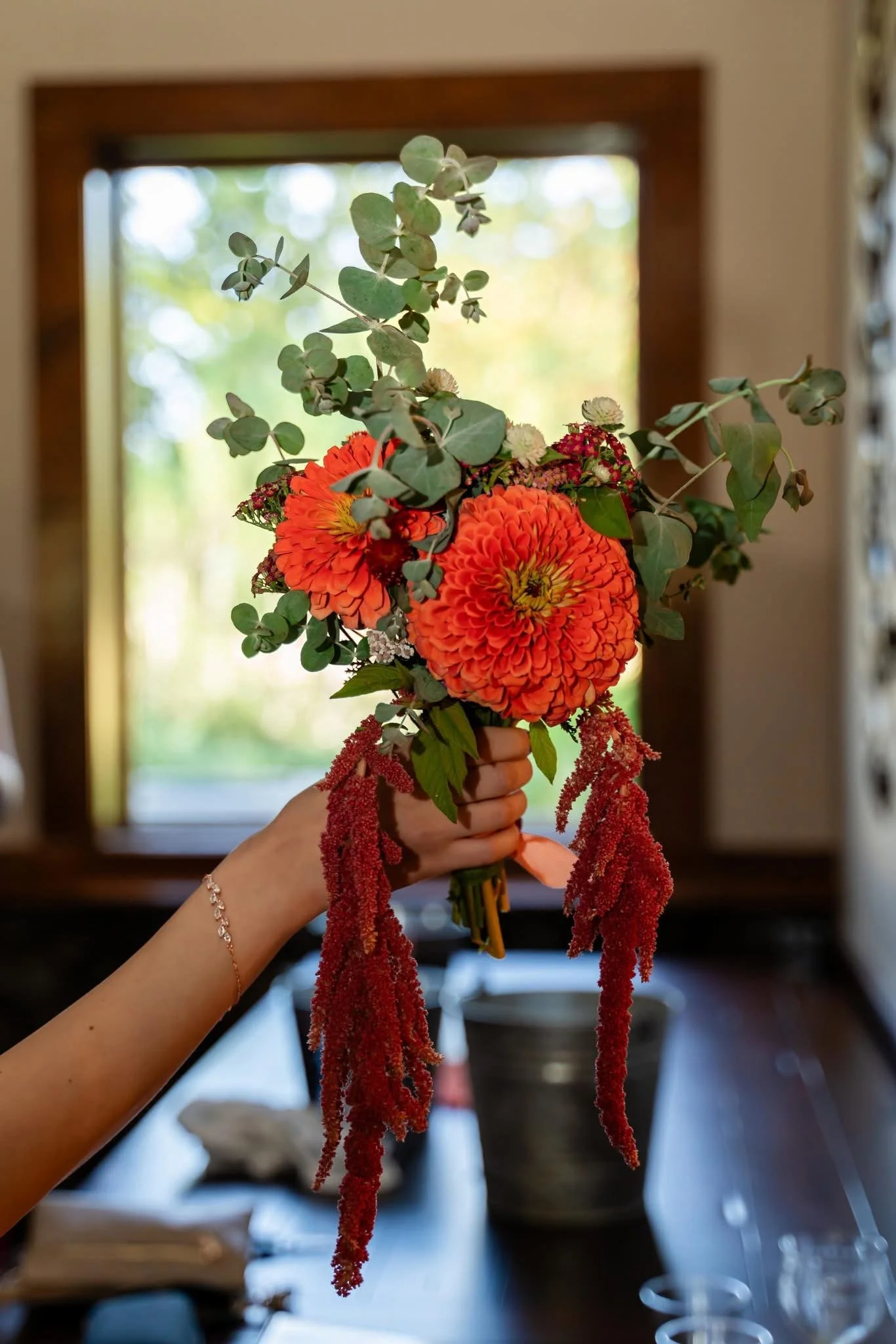 Person holding a bouquet of orange flowers, surrounded by green foliage, with a window in the background.