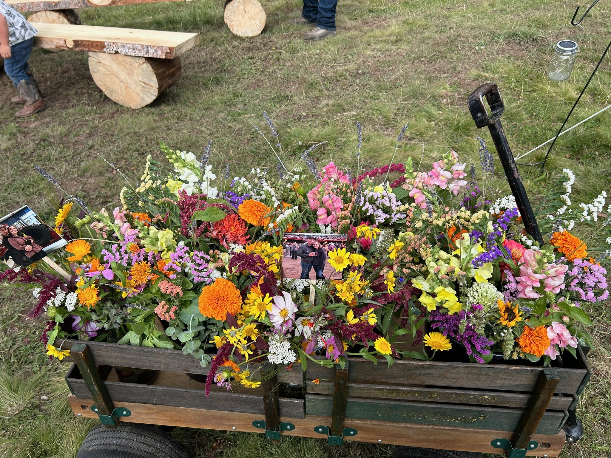 A cart filled with a colorful assortment of fresh flowers, including daisies, marigolds, snapdragons, and lavender, with a couple of photographs resting among the blooms. In the background, there are people and a grassy field.