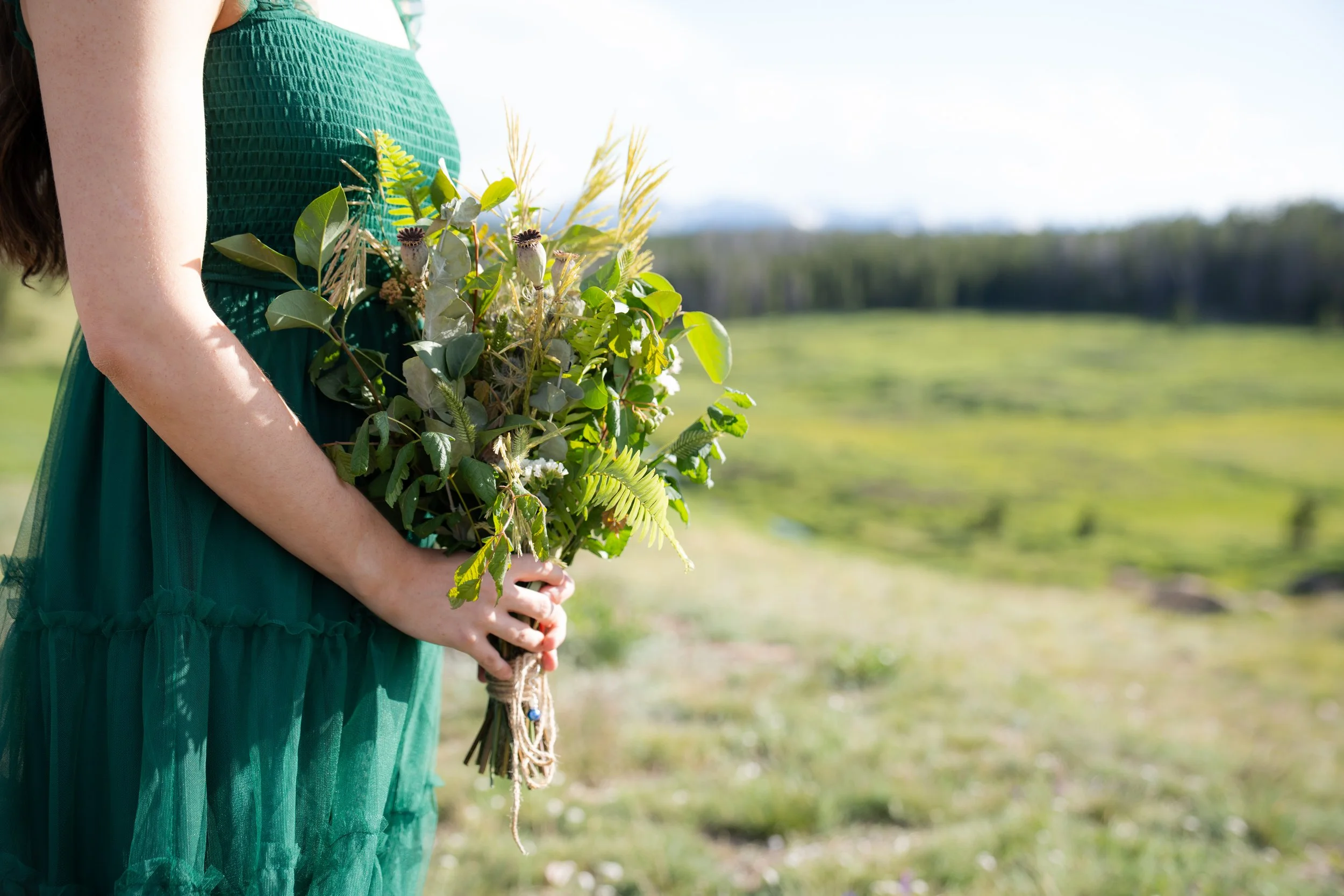 A woman wearing a green dress holding a bouquet of flowers and greenery in an outdoor setting with a grassy field and trees in the background.
