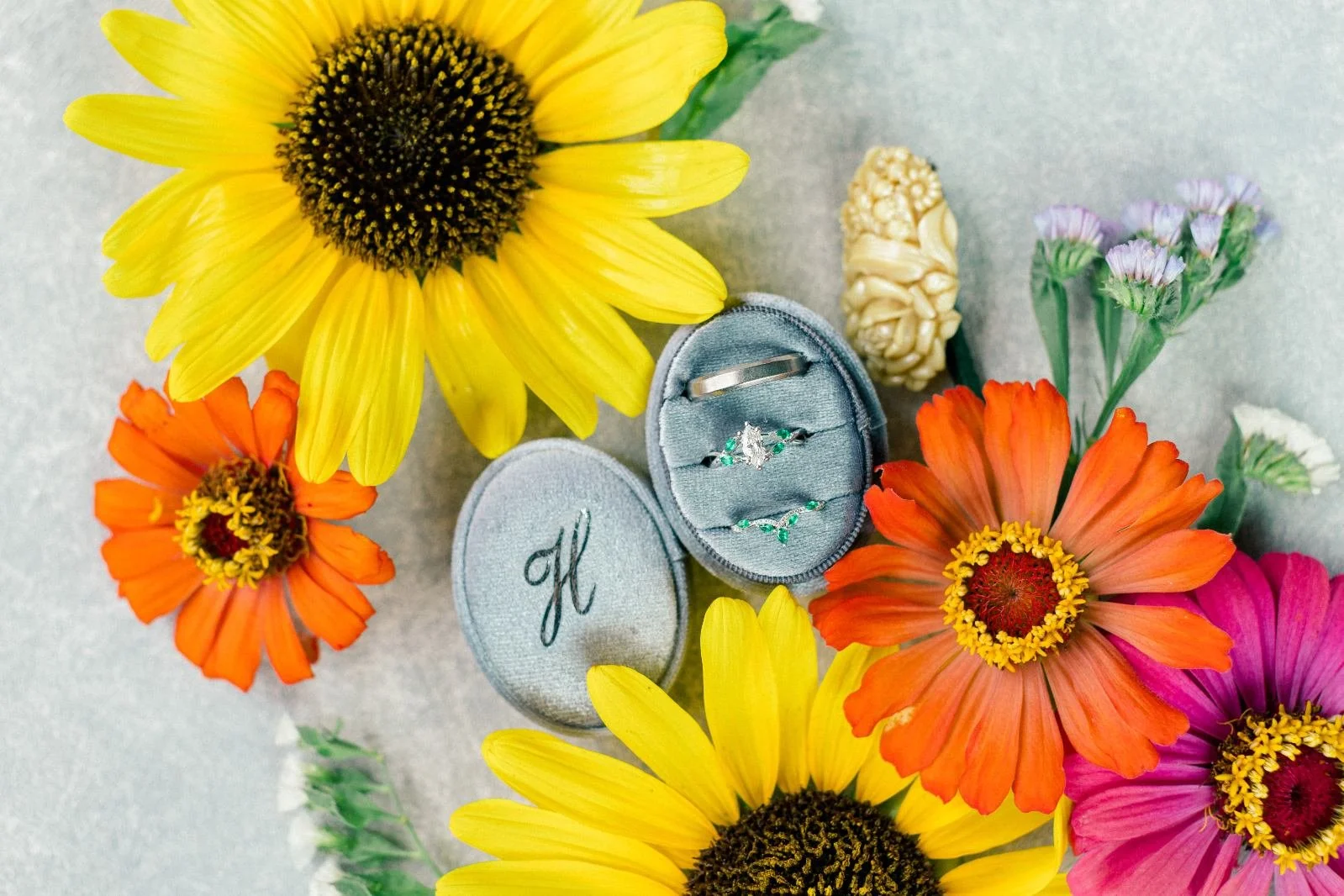 A sunflower and orange, pink, and purple flowers surround a velvet ring box with a diamond ring and emerald earrings, with a wedding band on top of the box.