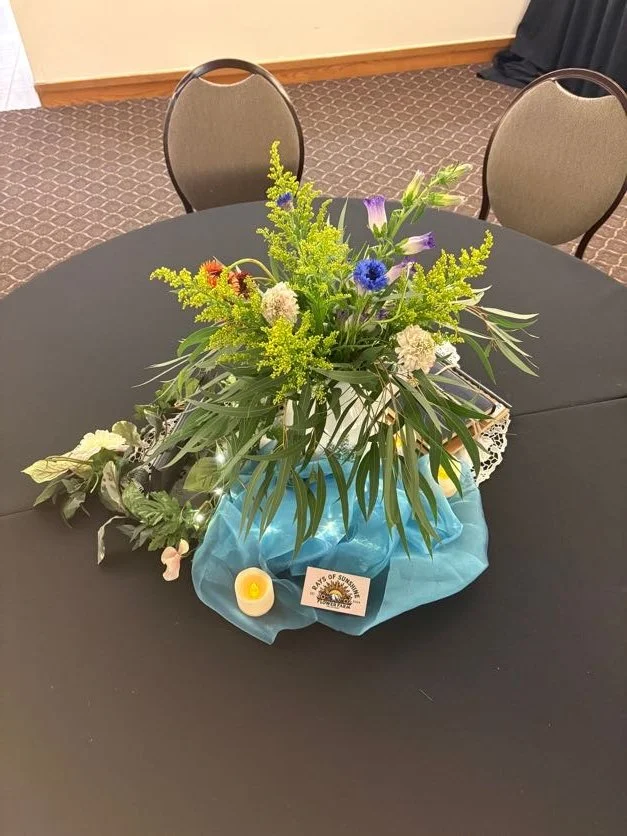 A floral centerpiece with various green leaves and colorful flowers on a black round table. The arrangement is decorated with a blue cloth, a small candle, and a card that says 'BANK OF SUMMER'.