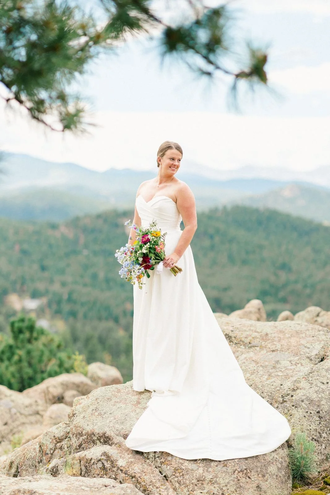 A bride in a white strapless wedding dress holding a colorful bouquet standing on a rock with a mountainous background and blue sky.
