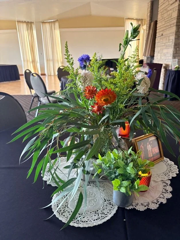 A floral centerpiece with various colorful flowers and green foliage on a doily-covered table, including a small framed photo and a red decorative item, in a room with curtains and chairs.