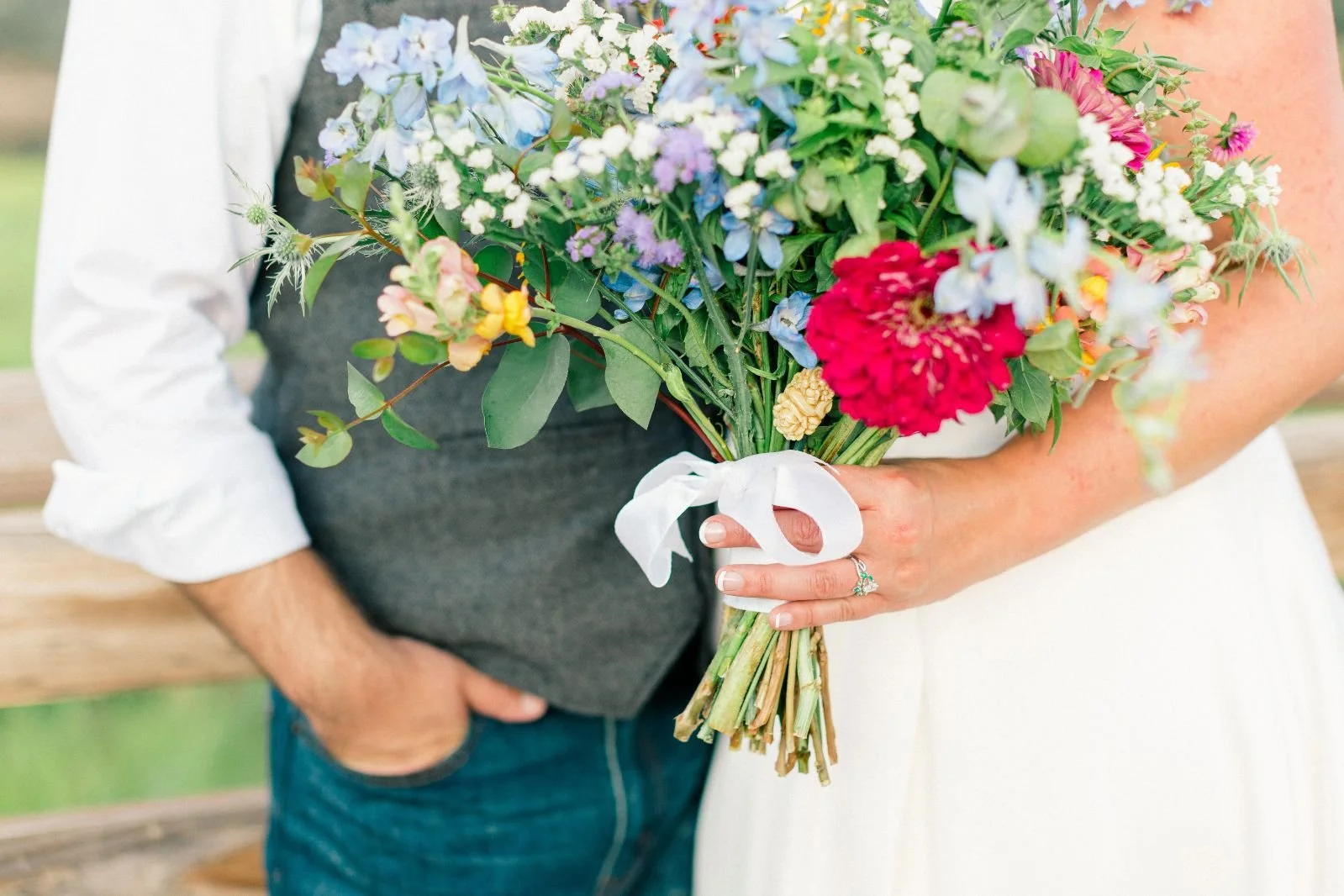 A woman holding a colorful bouquet of various flowers wrapped with a white ribbon, with a man standing beside her against a blurred outdoor background.