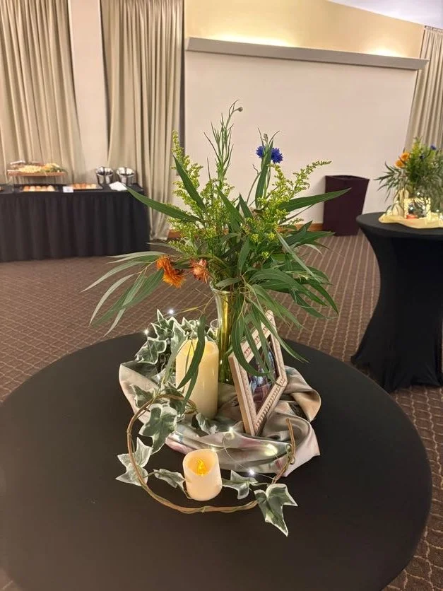 Centerpiece with green foliage, assorted flowers, a picture frame, candles, and decorative elements on a black table in an event space.