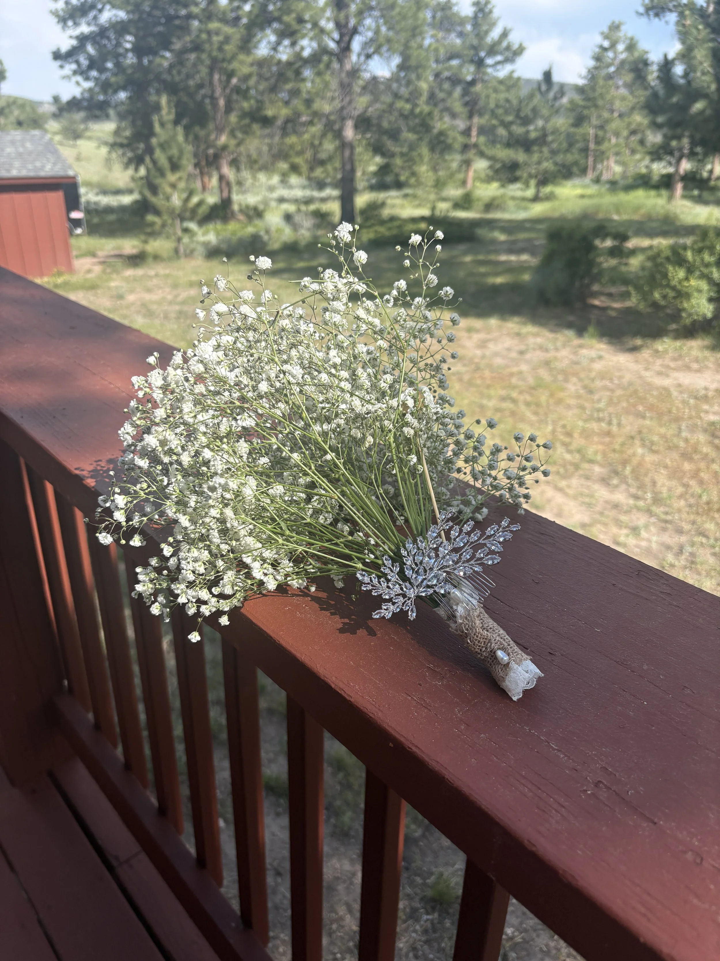 A bouquet of white baby's breath flowers with decorative silver leaves on a wooden porch railing, with a blurred background of green trees and a blue sky.