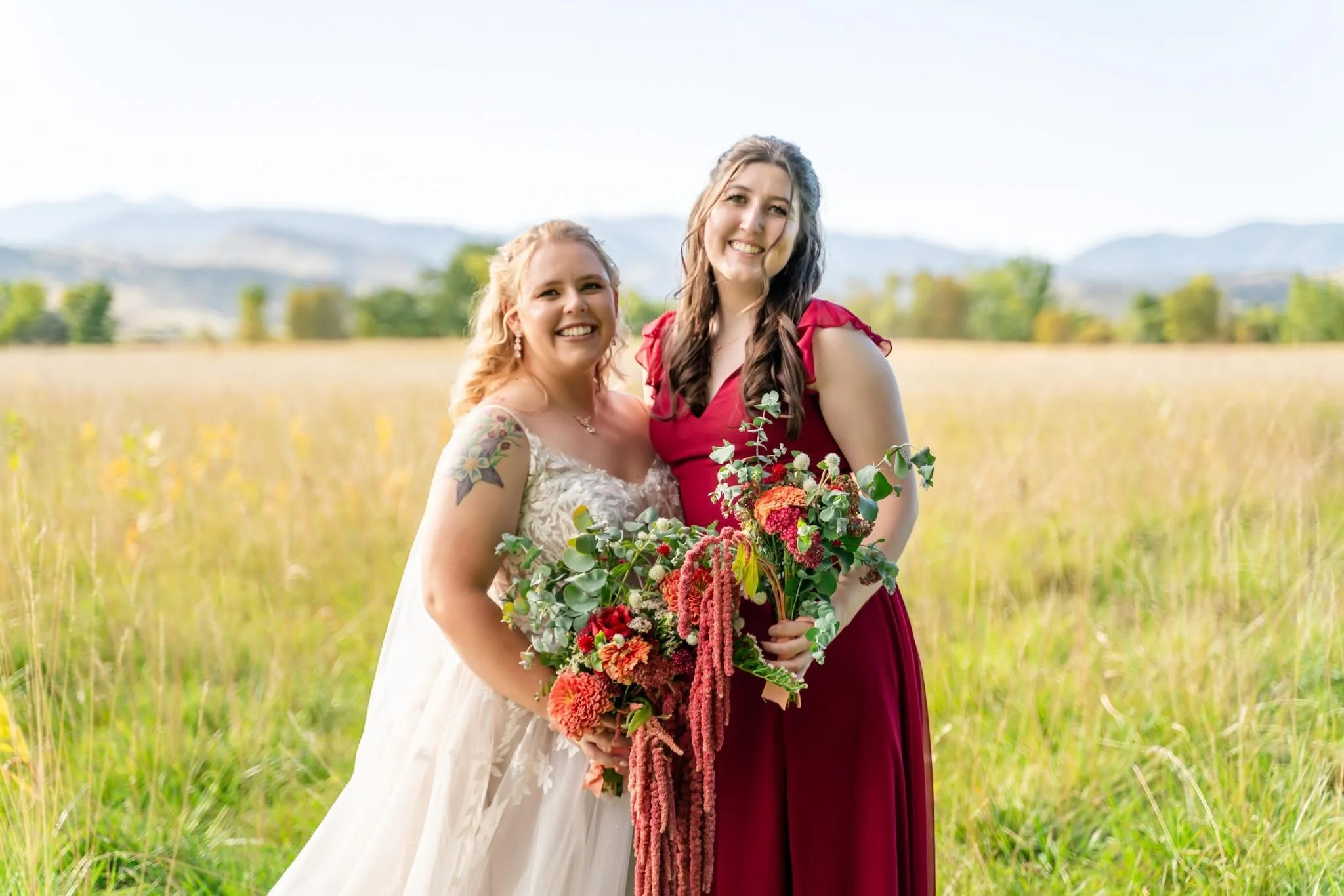 Two women standing in a grassy field, one wearing a white wedding dress and the other wearing a red dress, holding a bouquet of flowers, with a scenic landscape and mountains in the background.