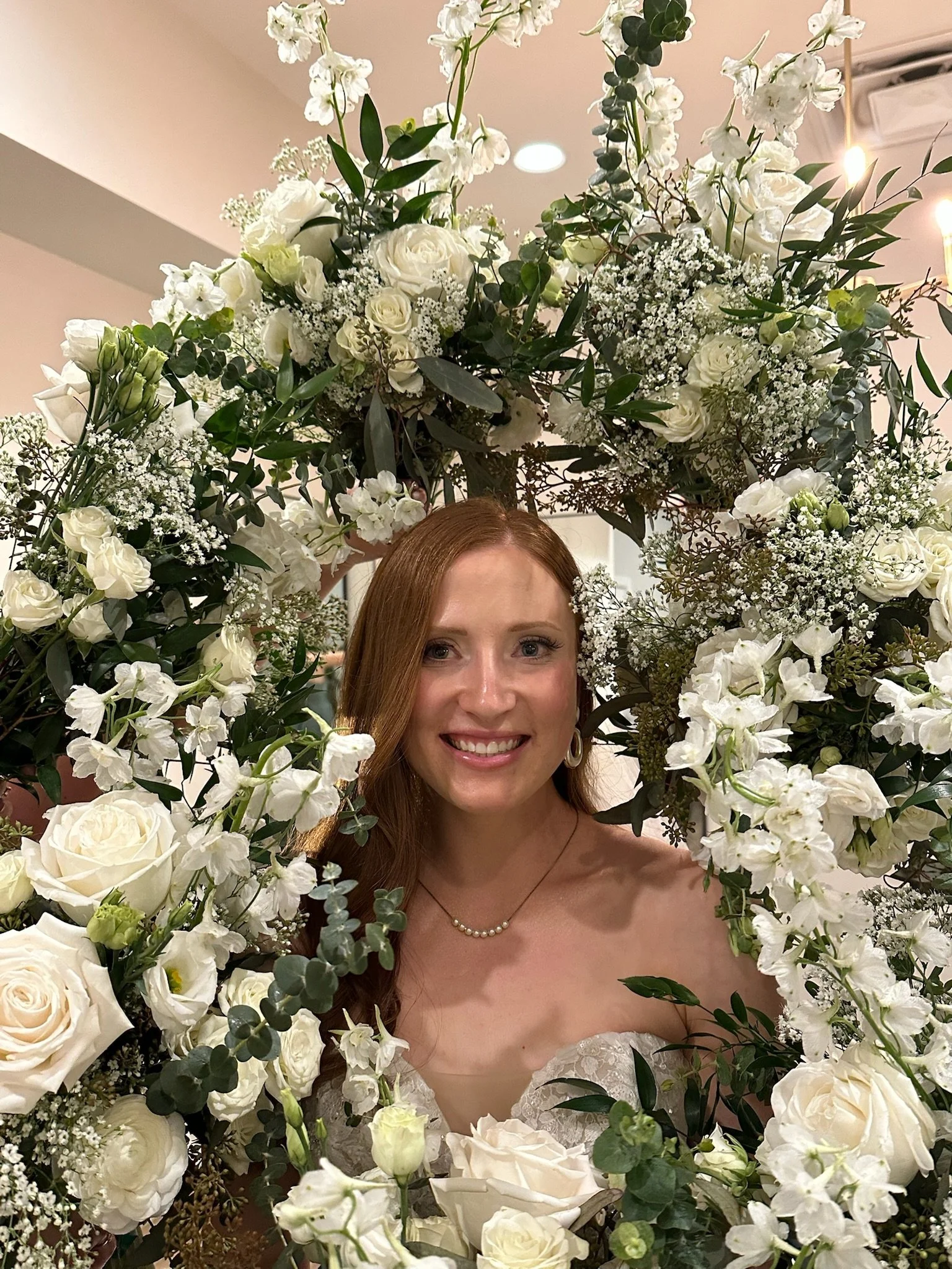 A woman with red hair is smiling while surrounded by a large, intricate floral arrangement of white roses, baby's breath, and greenery. Only her face and part of her shoulders are visible beneath the flowers.