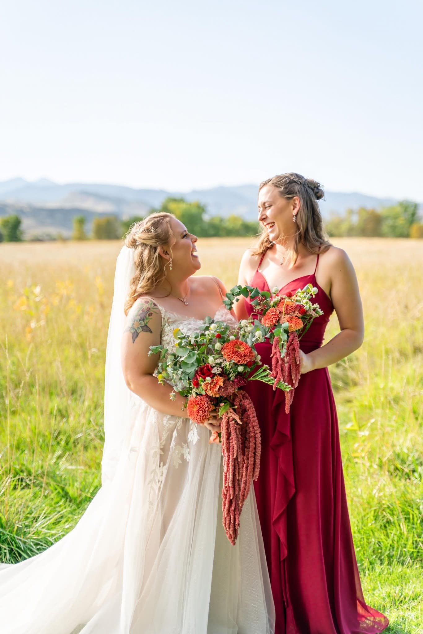 Two women, one in a wedding dress and one in a red gown, smiling and holding a bouquet of flowers in a field with mountains in the background.