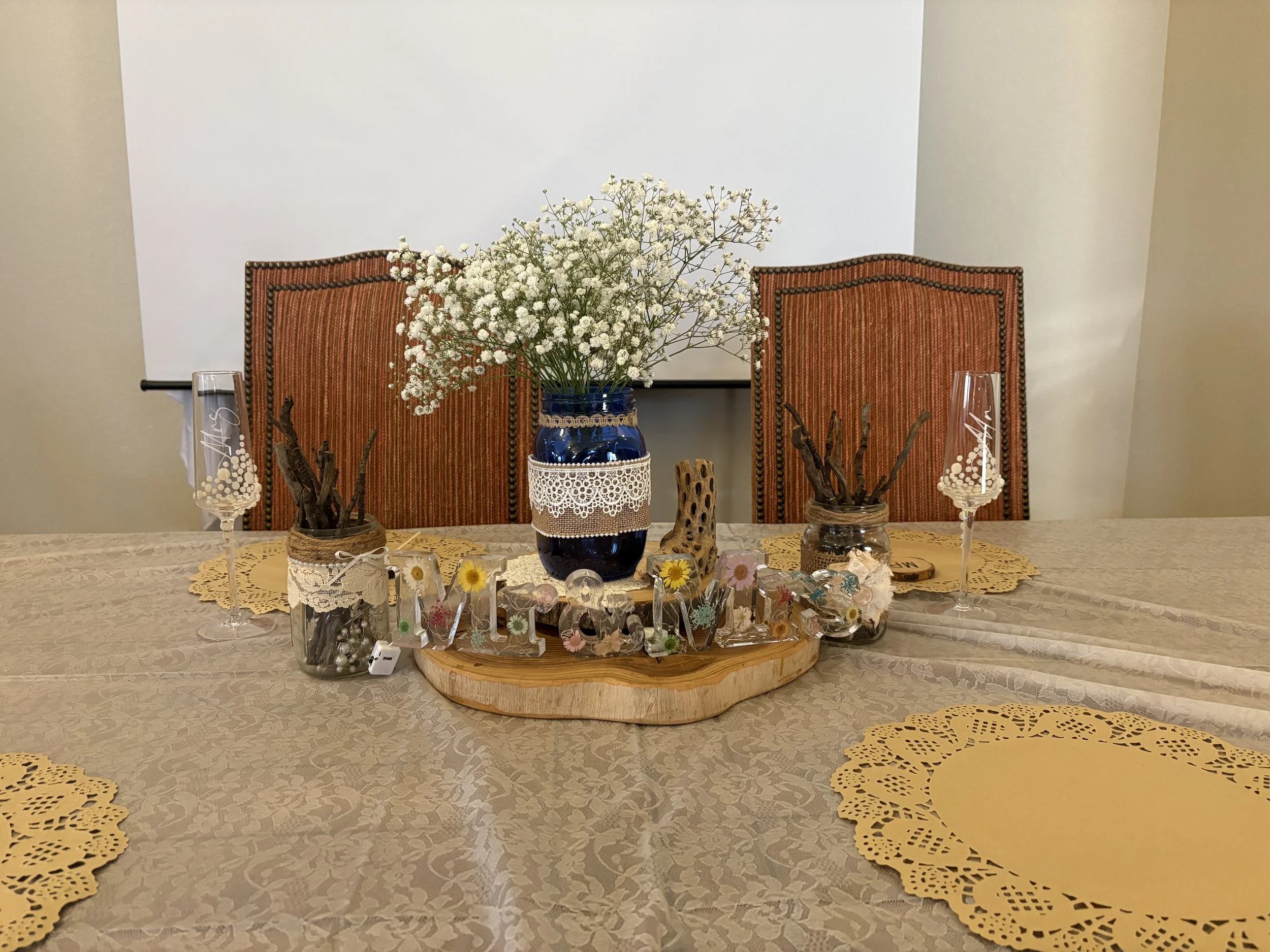 A dining table with a flower arrangement, decorated with yellow paper doilies, crystal glasses, candles, and small decorative items. Chairs are positioned behind the table against a white wall.