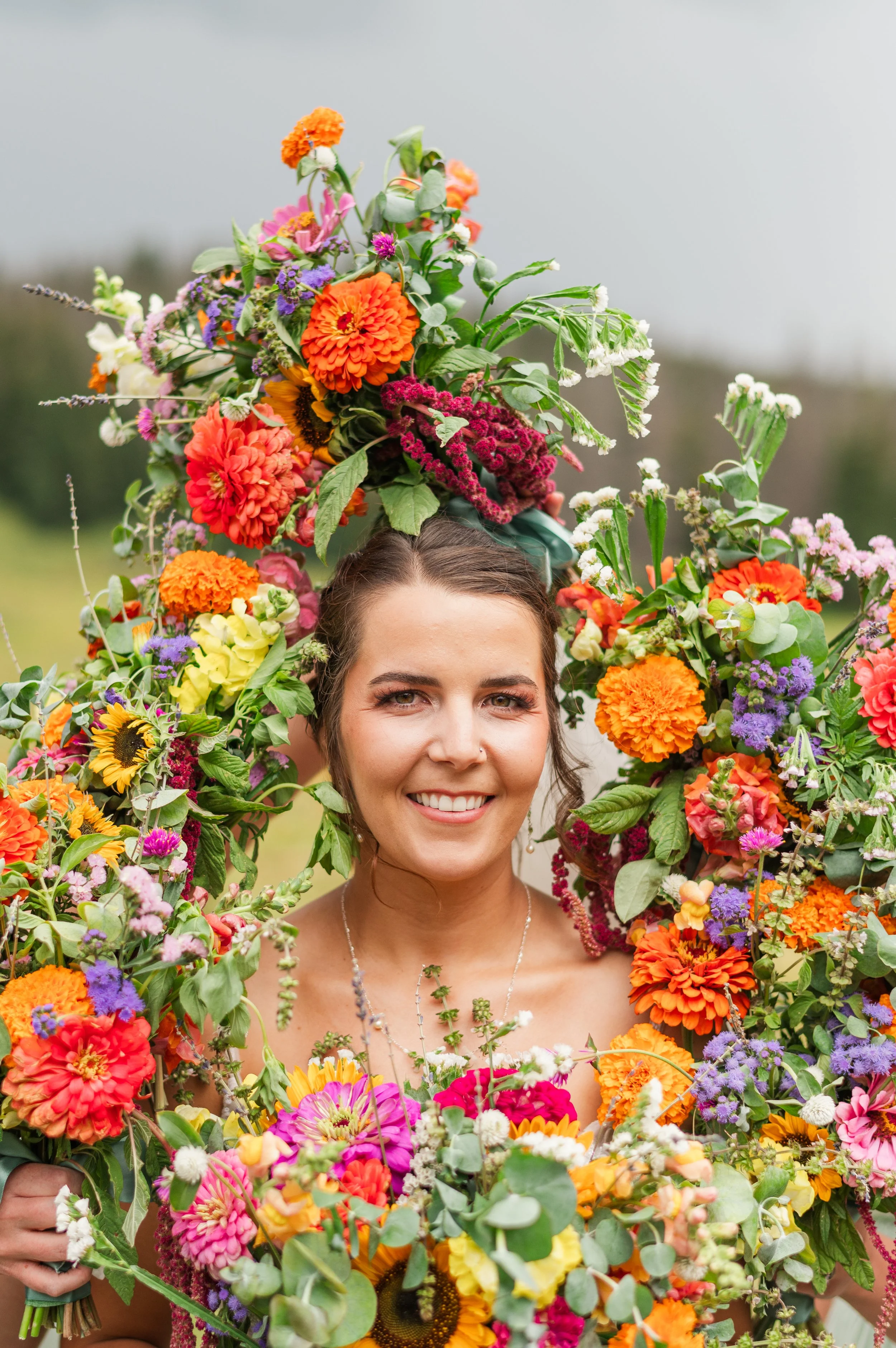 A woman smiling outdoors surrounded by colorful flowers and greenery, with a floral crown on her head.