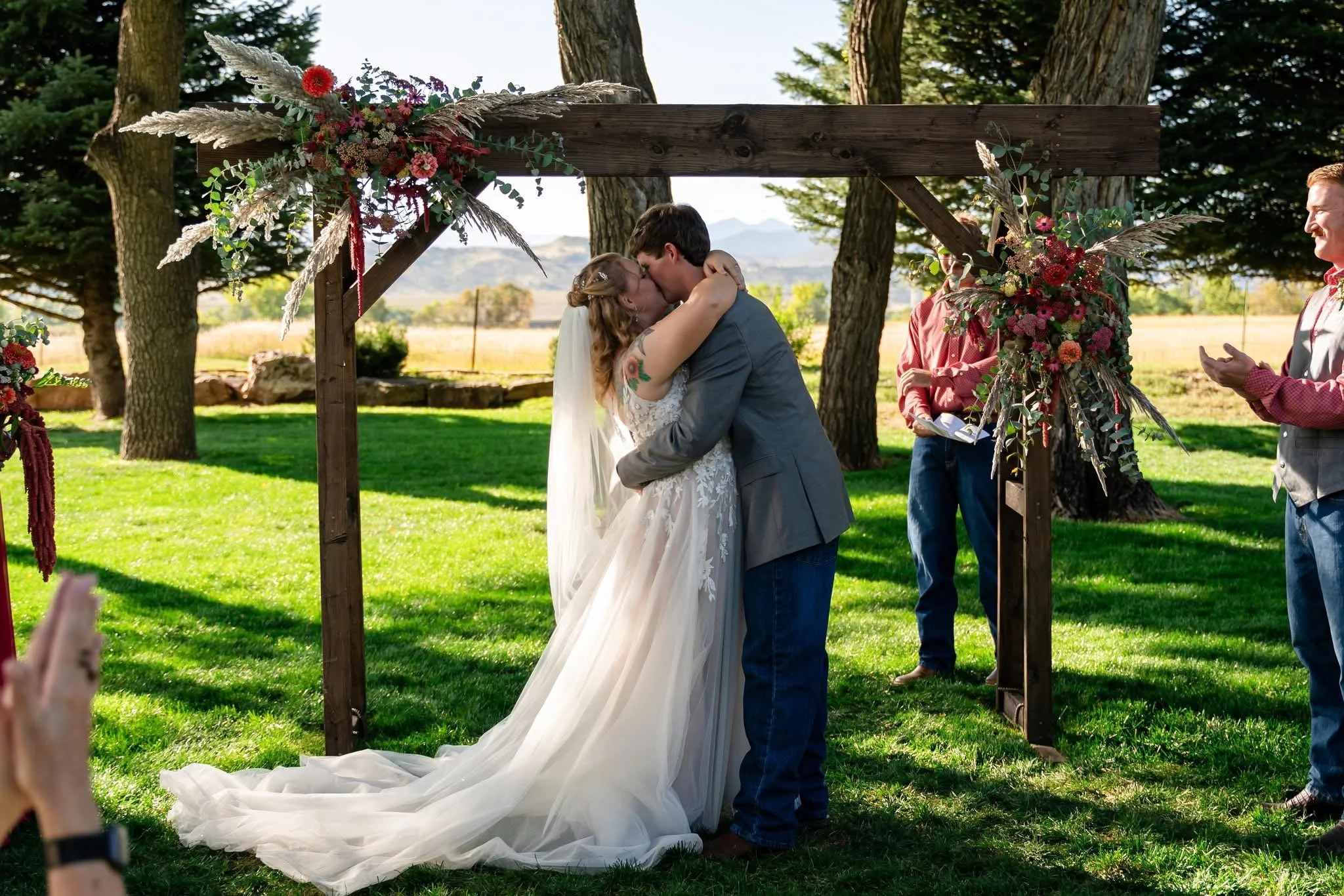 A couple kissing during their outdoor wedding ceremony, surrounded by friends and decorated with floral arrangements on a wooden arch, in a scenic natural setting with trees and mountains in the background.
