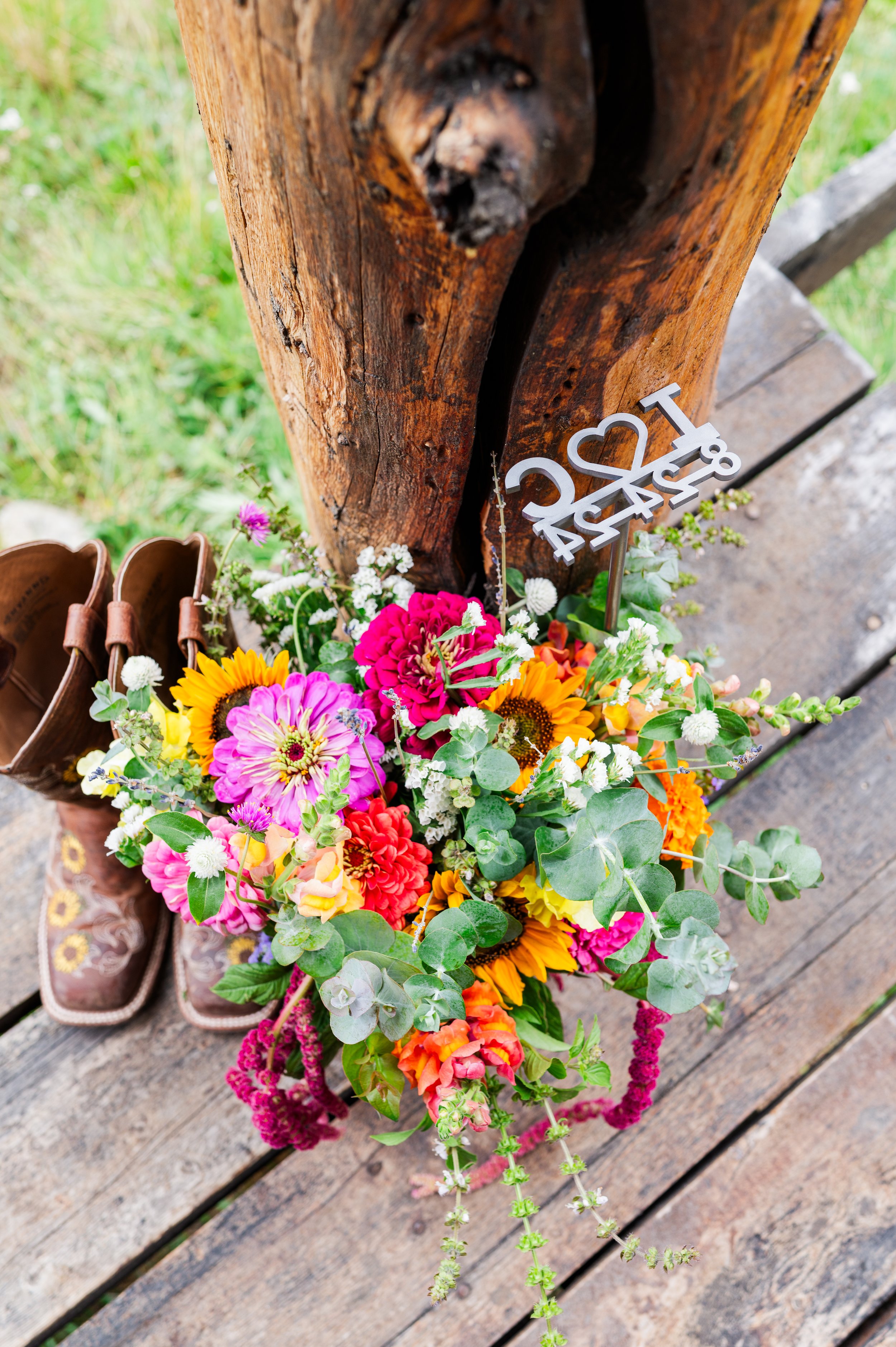 Colorful bouquet of flowers placed on a weathered wooden surface with a pair of cowboy boots nearby and a wooden post with a decorative metal sign.