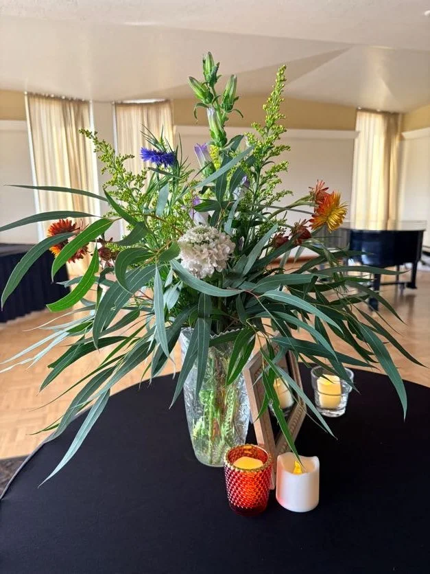 A floral arrangement in a glass vase on a black table with candles in red and white holders in front, set in a room with large windows and cream-colored curtains.