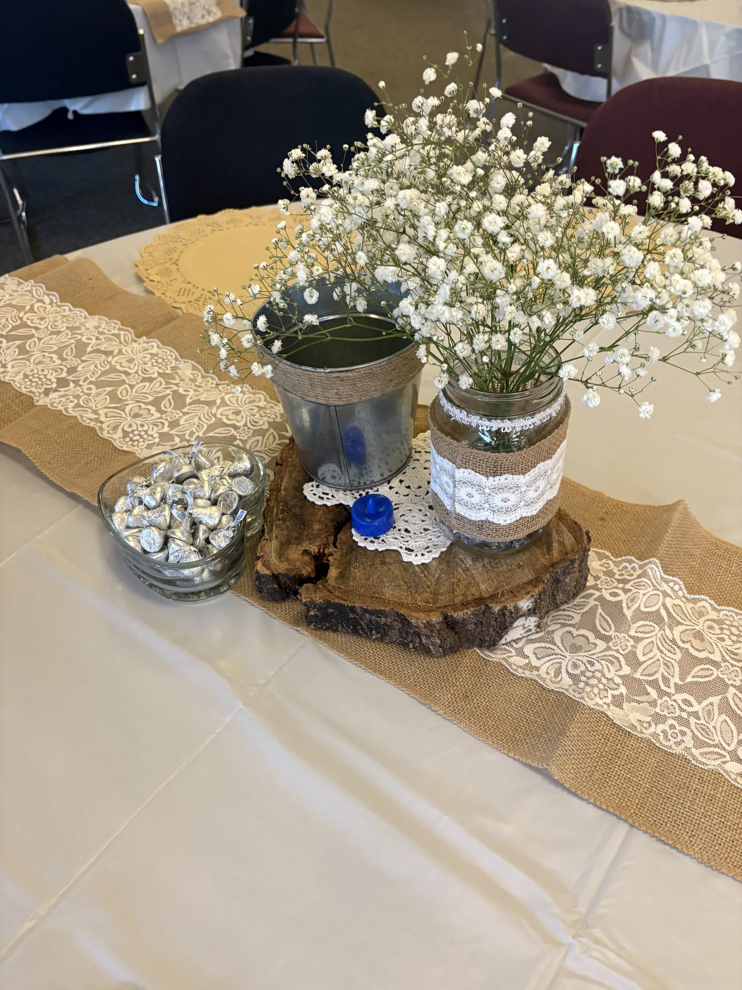 A wedding or event table centerpiece featuring a glass jar with lace and burlap decoration, filled with white baby's breath flowers, placed on a wooden slab. Surrounding it is a burlap table runner, a lace doily, a small blue candle, and a glass bowl