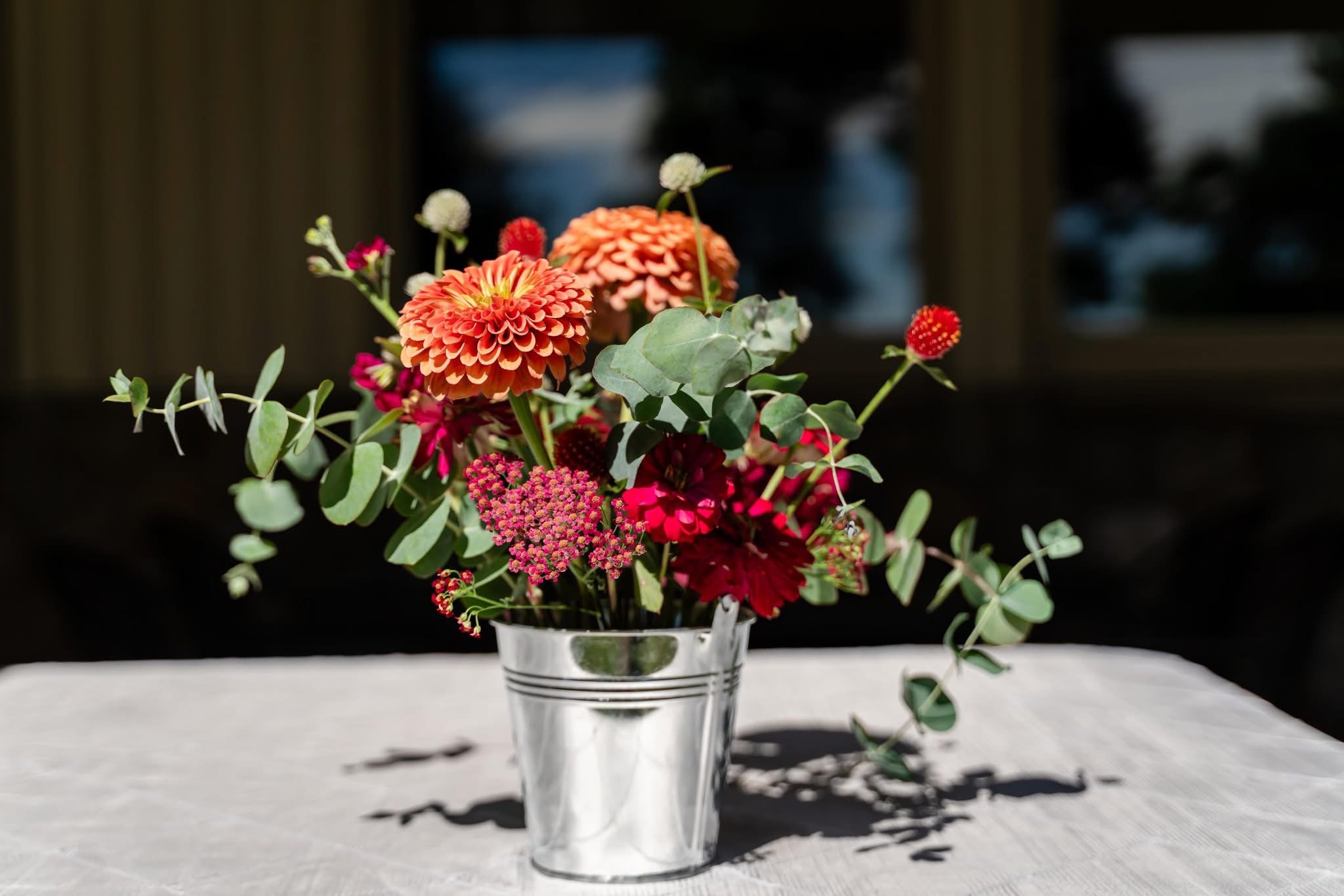 A bouquet of colorful flowers, including orange and red dahlias, pink and white flowers, and eucalyptus leaves, arranged in a glass vase on a white surface with shadows and reflections.