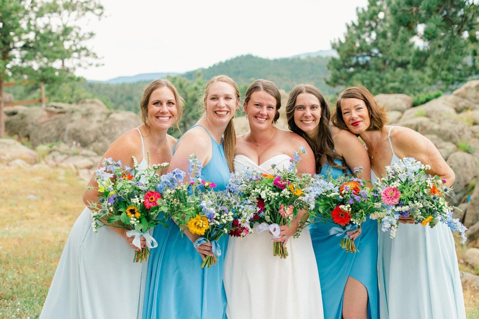 Group of women in wedding attire holding bouquets, standing outdoors with trees and rocks in the background.