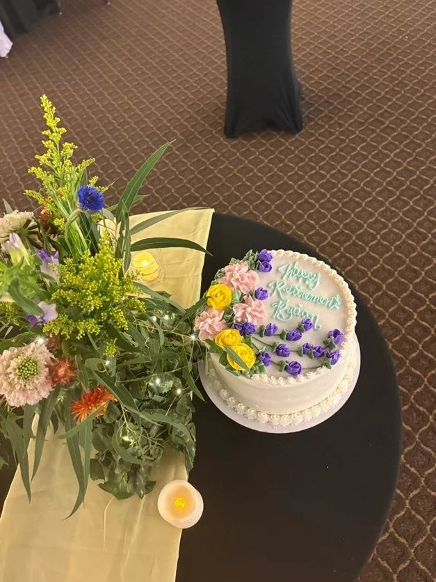 A birthday cake decorated with pink and purple flowers and yellow roses, with the message 'Happy Retirement Rhea' written on it. Next to the cake, there is a large floral arrangement with various colorful flowers and greenery on a black table with a 