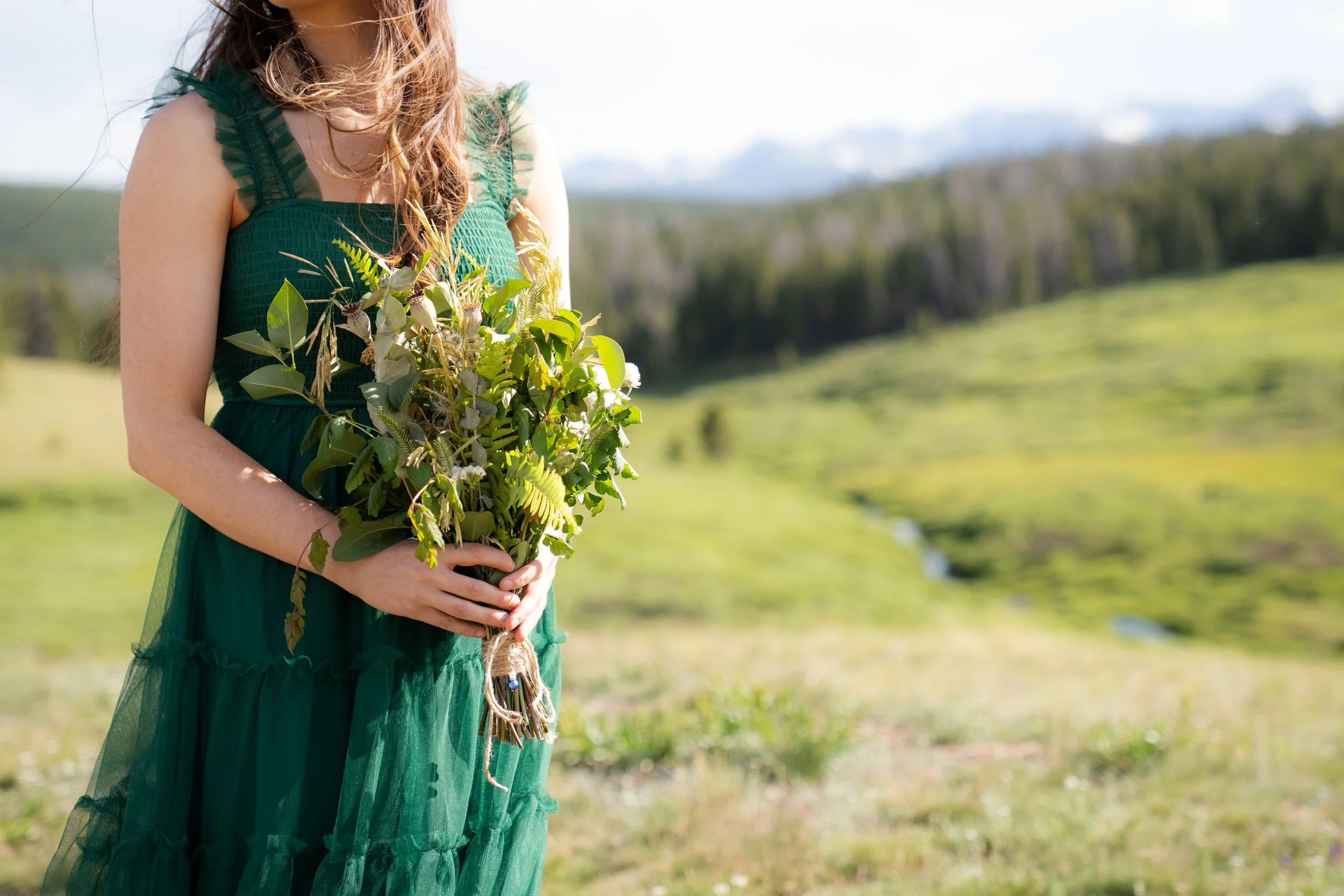 A woman in a green dress holding a bouquet of wildflowers standing outdoors in a grassy field with trees and mountains in the background.