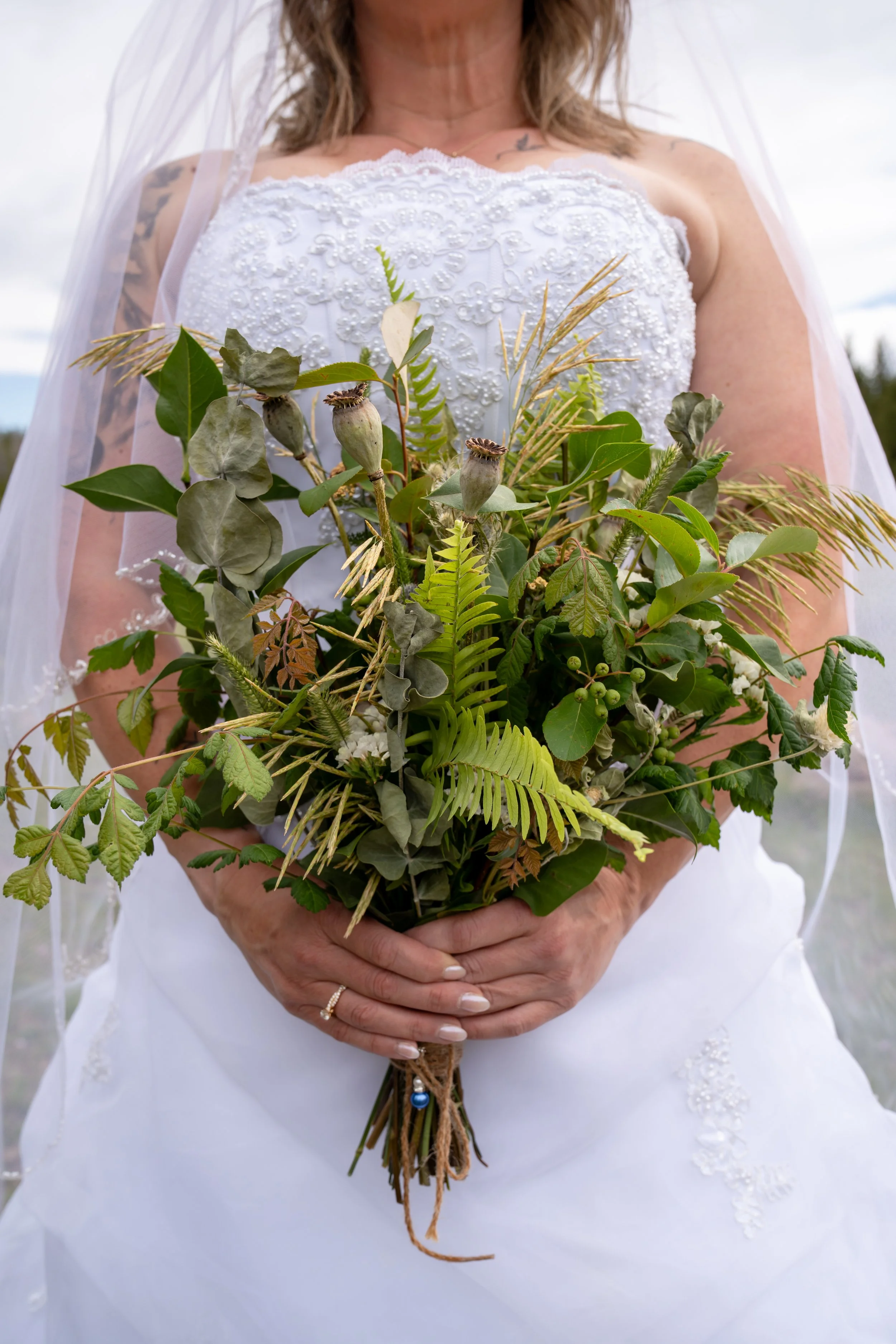 A woman wearing a white embroidered wedding dress, holding a bouquet of green foliage and seed pods, with her hands clasped in front of her.